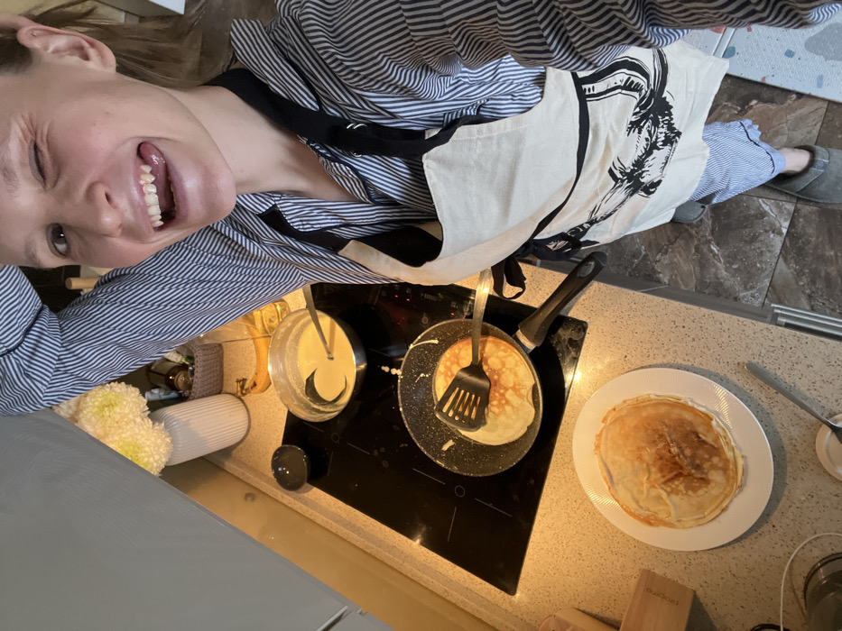 A female individual is depicted in a kitchen in Khimki, Russia, actively engaged in cooking. The woman, positioned at the right of the frame, is captured from the chest up, looking directly at the camera with a wide smile, a wink of her right eye, and her tongue slightly extended. She wears a blue and white striped long-sleeved shirt, a cream-colored apron featuring a dark graphic design, striped pajama bottoms, and grey slippers. Her right arm is raised above her head, with her hand near her hair.
The kitchen counter, a light-speckled surface, holds a black induction cooktop. On the front-left burner, a dark non-stick frying pan contains a pancake or crepe in the process of cooking, with a black slotted spatula resting upon it. To the right, a silver metal pot on the back-right burner holds a quantity of light-yellow batter, with a silver ladle submerged within it. A stack of several golden-brown cooked pancakes or crepes is presented on a white, ribbed-edge round plate situated on the countertop to the left of the stovetop.
Further objects on the counter include a small dark bowl to the left of the silver pot, a white ribbed vase holding yellow-white chrysanthemums, a woven basket, a dark bottle, a single yellow banana, and a clear bottle of cooking oil positioned behind the cooking area. A wooden block is visible on the far left. The floor beneath the woman's feet consists of dark, patterned tiles. A light grey overhead cabinet is partially visible in the upper left corner of the scene.