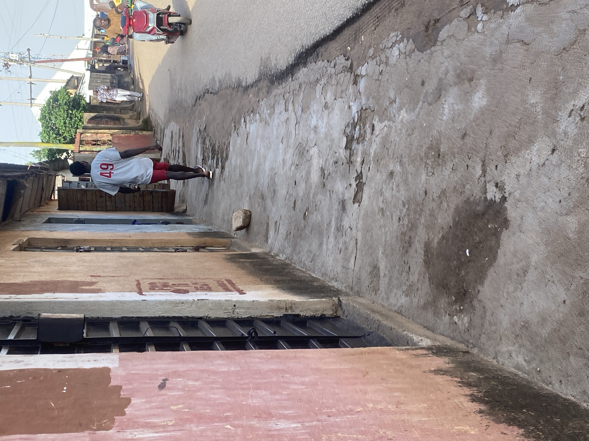 The image depicts a street scene in Jos, Nigeria, viewed from a low angle looking down a cracked concrete sidewalk. On the left, a building with a salmon-pink facade and barred windows dominates the foreground. The sidewalk itself is worn and shows patches of dark discoloration, possibly from water or oil stains. A young man wearing a white t-shirt with the number "49" in red and red shorts walks away from the camera down the sidewalk. Further down the street, a red motorbike is being ridden by a man wearing a yellow shirt. The background shows more buildings, some with corrugated metal roofs, and overhead power lines. The overall impression is of a typical urban street environment in Nigeria, characterized by weathered infrastructure and pedestrian and vehicular activity.
