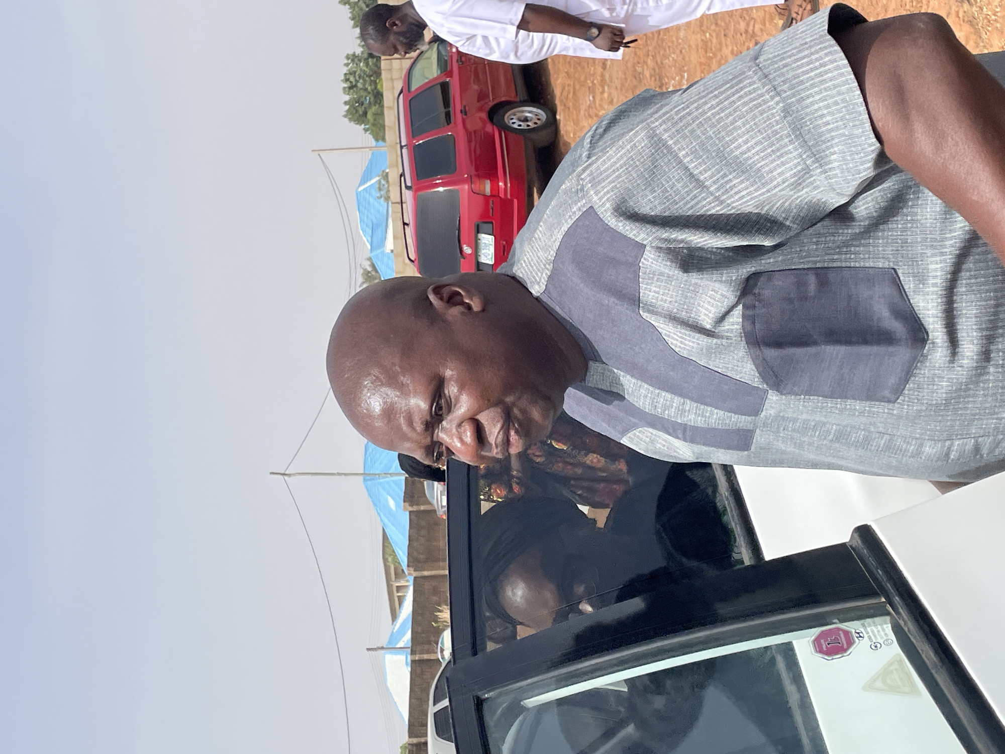 A man with a bald head, wearing a grey shirt with a dark grey pocket, is seen looking into the open driver's side window of a white vehicle. Another person is in the driver's seat. Outside the car, there is a red vehicle. In the background, there are blue tents and another man in white clothing. The scene appears to be located in Bar Jirgi Summa, Nigeria.
