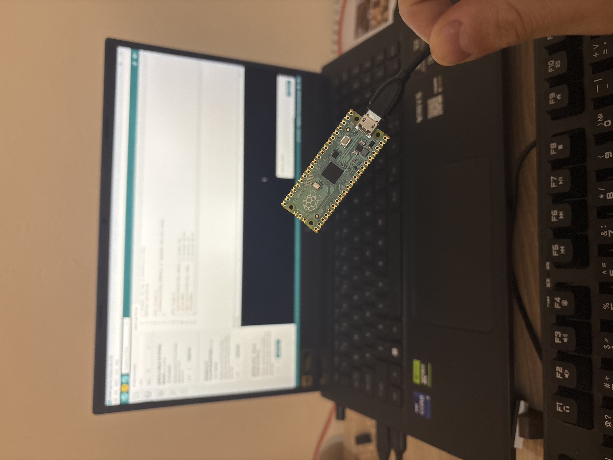 The image shows a person holding a Raspberry Pi Pico microcontroller board connected to a USB cable. The board is positioned in front of a laptop screen displaying what appears to be the Arduino IDE. A black keyboard is visible to the right of the laptop. The image provides no information that indicates a location of this scene in the city of Plovdiv, Bulgaria.