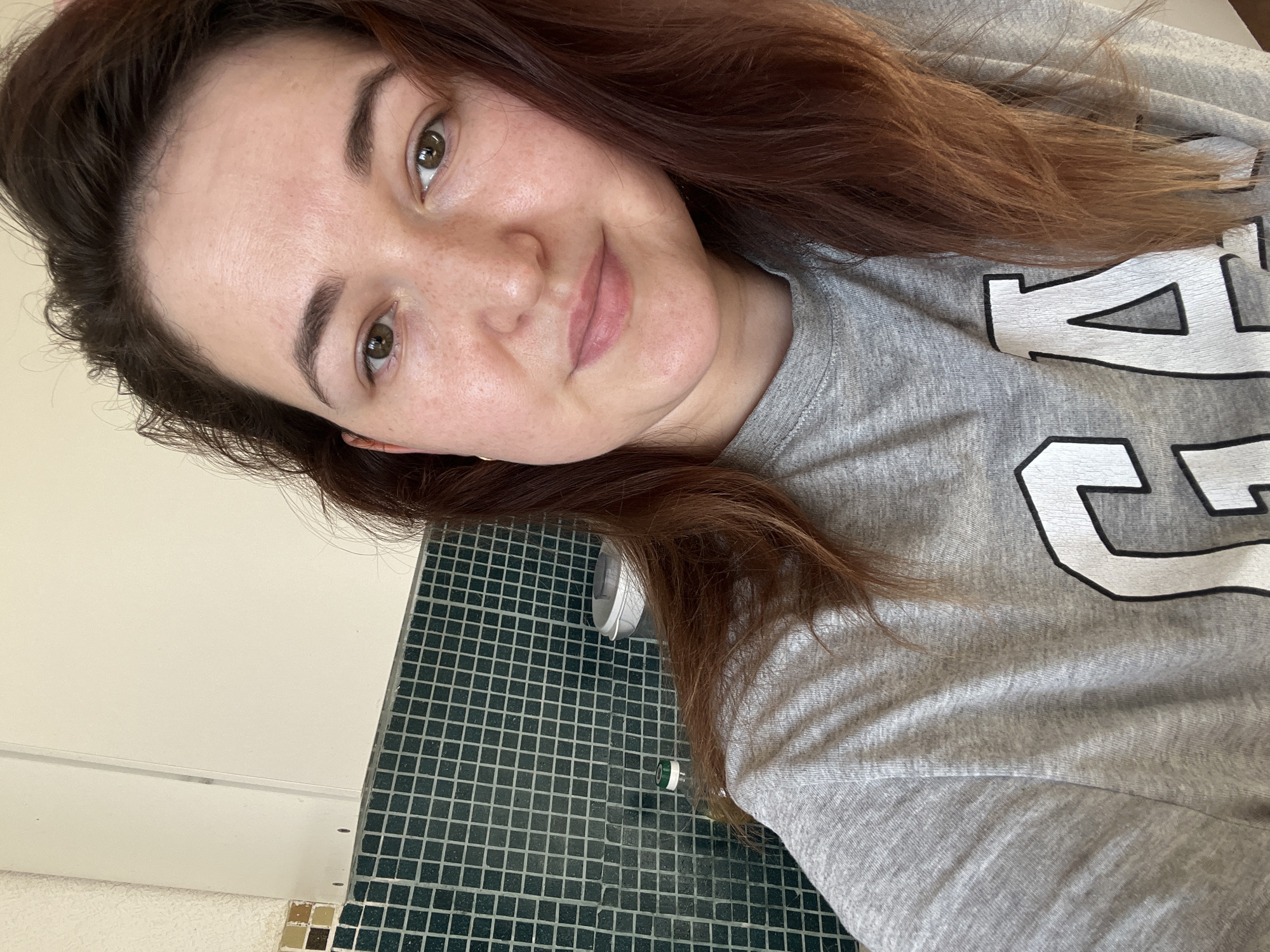 A young woman with reddish-brown hair and light skin with freckles is taking a selfie. She is wearing a gray sweatshirt with a large white and black design that appears to be a letter or logo. Her head is tilted to the side, and she is looking directly at the camera with a slight smile. Her eyes are a warm brown.
The background of the image shows a wall with a tiled surface. The tiles are small, square, and dark green with light gray grout. There is also a sliver of a white surface visible on the left. In the lower part of the image, on the tiled surface, a white cap and a clear plastic bottle with a green cap are visible.
The lighting suggests it is daytime, but no specific time can be determined. The setting appears to be indoors, possibly a bathroom or kitchen area, given the tiling. The woman's expression is relaxed and casual. No specific activity is taking place other than the selfie itself. The visible text on her sweatshirt is a large graphic element that is partially obscured by her hair and the frame of the image.