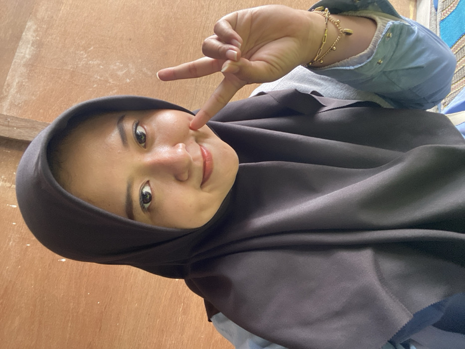A young woman wearing a dark hijab smiles at the camera, her head tilted slightly. Her right index finger touches her cheek, while her hand displays a peace sign. She wears a light blue button-down shirt and a gold bracelet. The background appears to be a wooden wall or panel. The image is taken at a low angle, with the woman looking down into the camera.