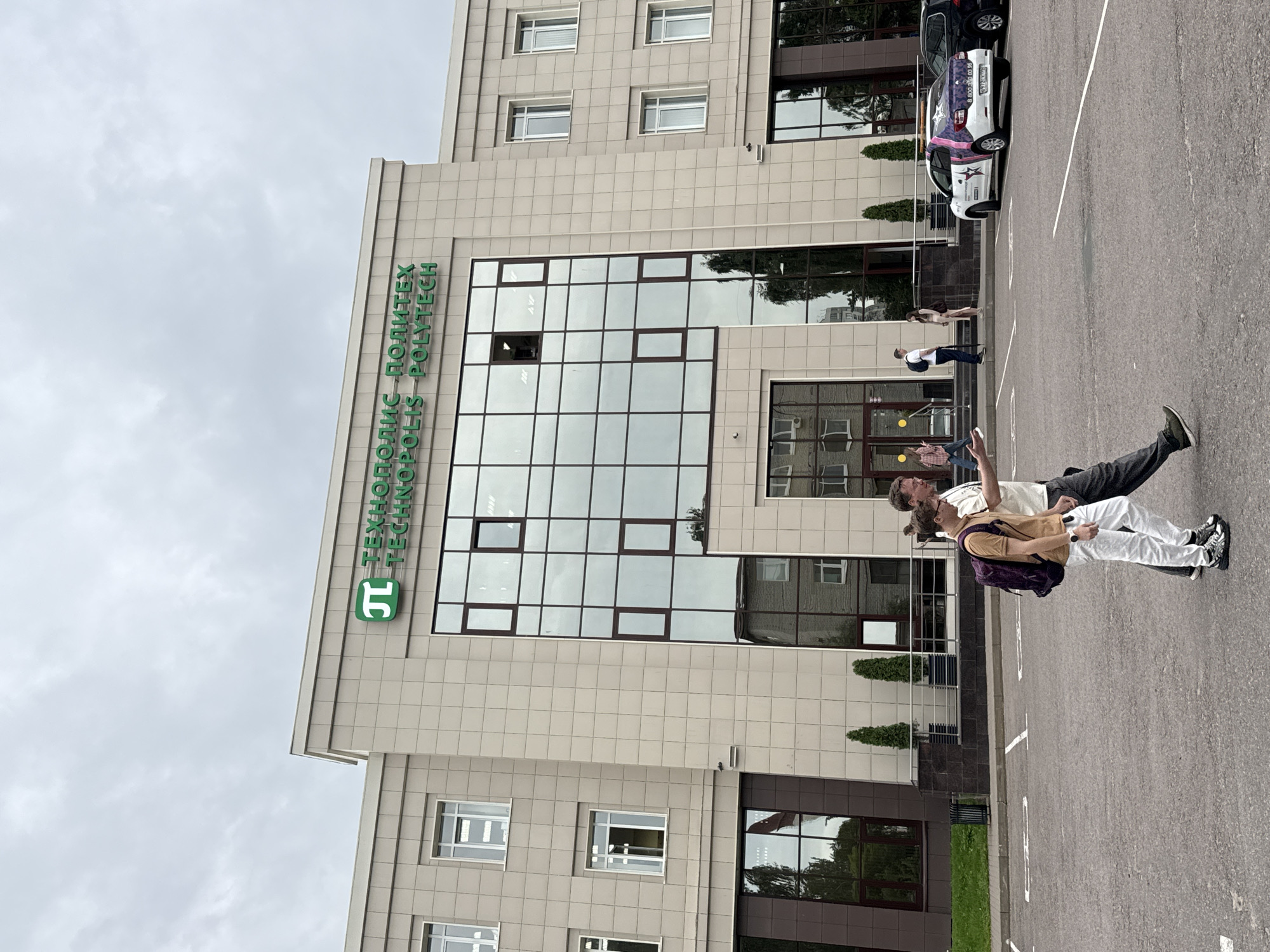 The image shows the facade of a modern, multi-story building with beige tiled walls and large, reflective windows. A green logo and the text "ТЕХНОПОЛИС ПОЛИТЕХ" and "TECHNOPOLIS POLYTECH" are visible at the top of the building. In the foreground, two young men are walking across a paved area that appears to be a parking lot. One man is wearing a light brown t-shirt and white pants with a dark backpack, while the other is wearing a light-colored t-shirt and dark pants. To the right of the men, a white car with a colorful graphic on its rear is parked. Further in the background, other cars and people are visible near the entrance of the building. The sky is overcast. The scene is likely located in Ladozhskoe Ozero, Russia, given the provided context.