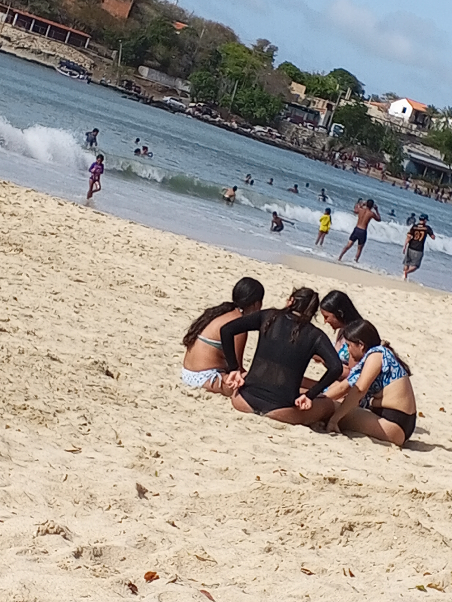 The image shows a beach scene with several people in Pedro González, Venezuela. In the foreground, four young women are sitting on the sand in a close group, possibly interacting or engaged in a conversation. Their attire includes swimsuits and a long-sleeved dark top. Behind them, the beach extends into the ocean where people are swimming and playing in the water. The water has small waves, and there are several people of various ages and genders in the water, some closer to the shore than others. Houses and vegetation are visible in the background along the shoreline. The weather seems sunny and clear.