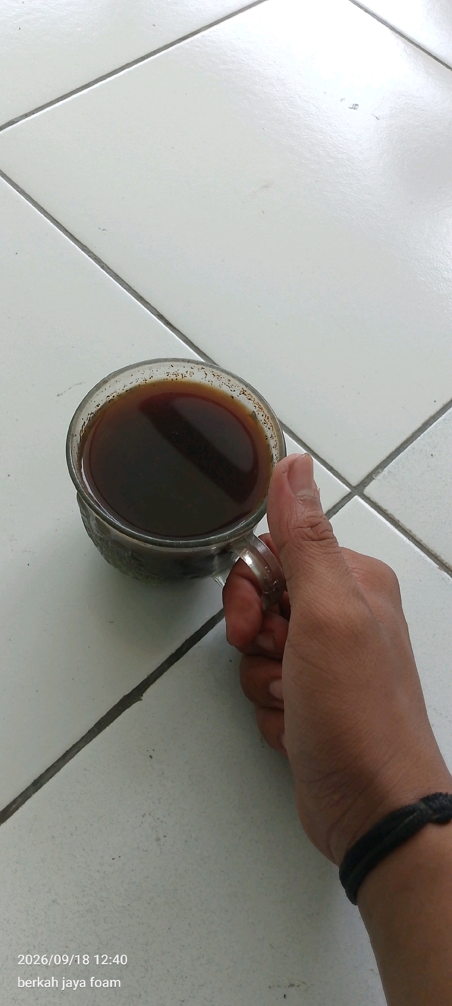 A hand is holding a glass of dark liquid, likely coffee, against a tiled background. The tiles are white and arranged in a grid pattern with dark grout lines. The hand has a black woven bracelet on the wrist. The timestamp "2026/09/18 12:40" and the text "berkah jaya foam" are visible in the lower left corner. The lighting suggests an indoor or shaded outdoor environment. The scene appears to be a casual moment, possibly at home or a similar personal space.