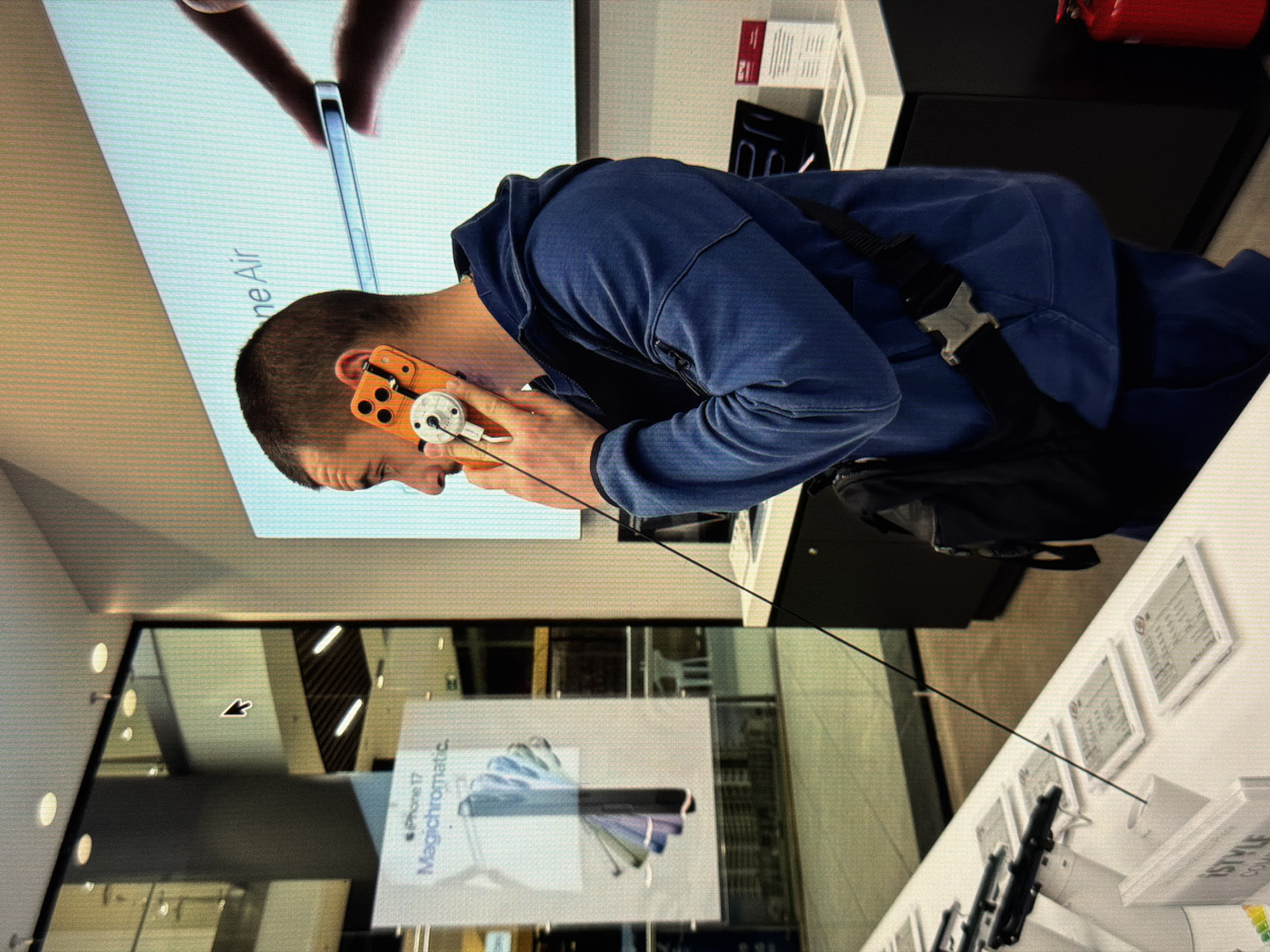 The image shows a man in a blue hoodie standing in front of a display of phones. He is holding an orange and silver device up to his ear. A black bag with a silver buckle is slung across his body. The background includes a display for "iPhone Air" and "iPhone 17 Magicchromatic". The location is a store in the city of Unknown, Unknown.