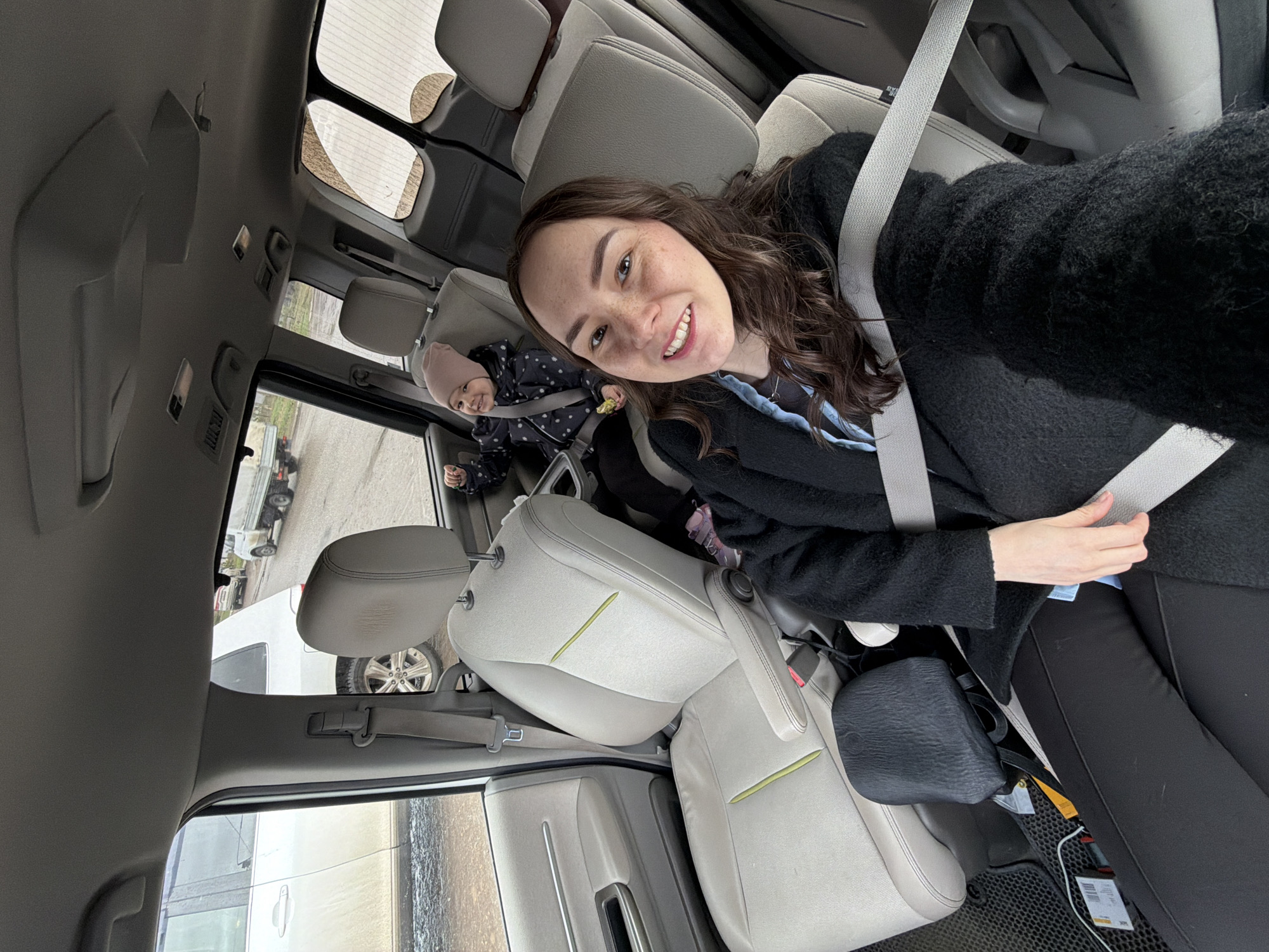 The image shows the interior of a vehicle. A woman with brown hair is in the passenger seat, wearing a black coat and a seatbelt. A young child, possibly a toddler, is seated behind her, also secured with a seatbelt and wearing a pink hat and patterned jacket. The vehicle appears to be stationary, as another vehicle is visible through the windows. The car's interior includes light-colored seats and interior trim. No specific location in Chaikovsky, Russia, is discernible from the image.