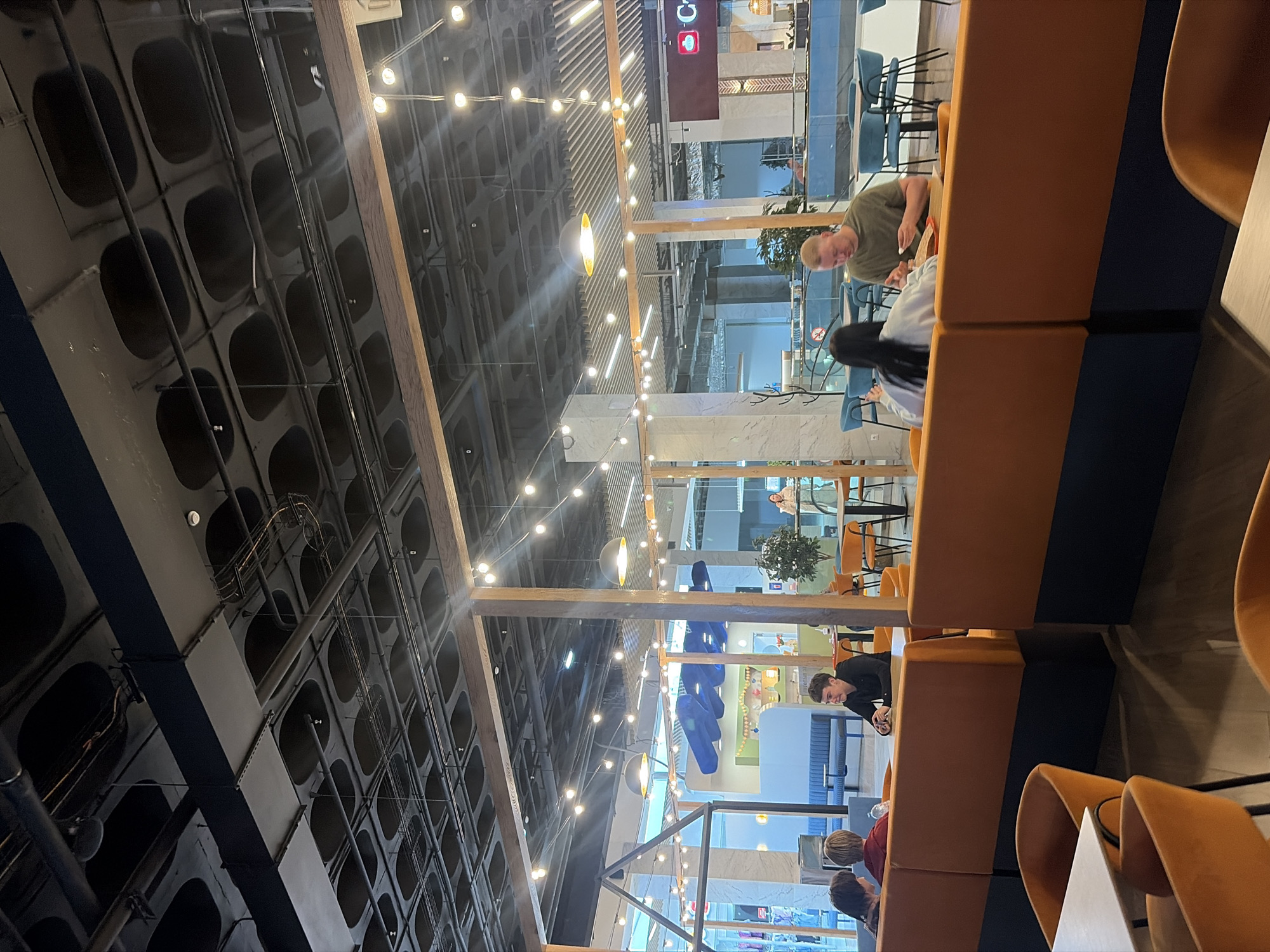 The image captures a vibrant, modern cafe or food court within a larger building, likely a shopping mall, in Izhevsk, Russia. The scene is brightly lit by numerous string lights draped across wooden beams that traverse the space, alongside recessed and linear lighting fixtures.
The high ceiling features an industrial design with a dark, possibly concrete, surface characterized by an array of recessed oval or circular patterns. Exposed pipes and ventilation ducts are visible, adding to the utilitarian aesthetic.
Seating arrangements consist primarily of upholstered booths in contrasting orange and dark blue colors, paired with light-colored tables. Additional chairs, some blue and others orange, are also present.
Several individuals are seated at tables and in booths, engaged in casual dining or conversation. In the foreground, a man with light-colored hair wearing a green t-shirt is visible from the chest up, looking down at his table. Opposite him, another person with dark hair is also looking downwards. Further into the scene, a man in a black long-sleeved shirt is seen leaning forward at a table. Other patrons are partially visible throughout the establishment.
In the background, large glass partitions or windows reveal a broader, brightly lit public area, suggesting the cafe is part of a larger commercial complex. A visible red sign with white lettering clearly displays "COFFEE" along with a coffee cup logo, indicating the type of establishment. The overall atmosphere appears casual and lively, suggesting daytime hours given the ambient light.