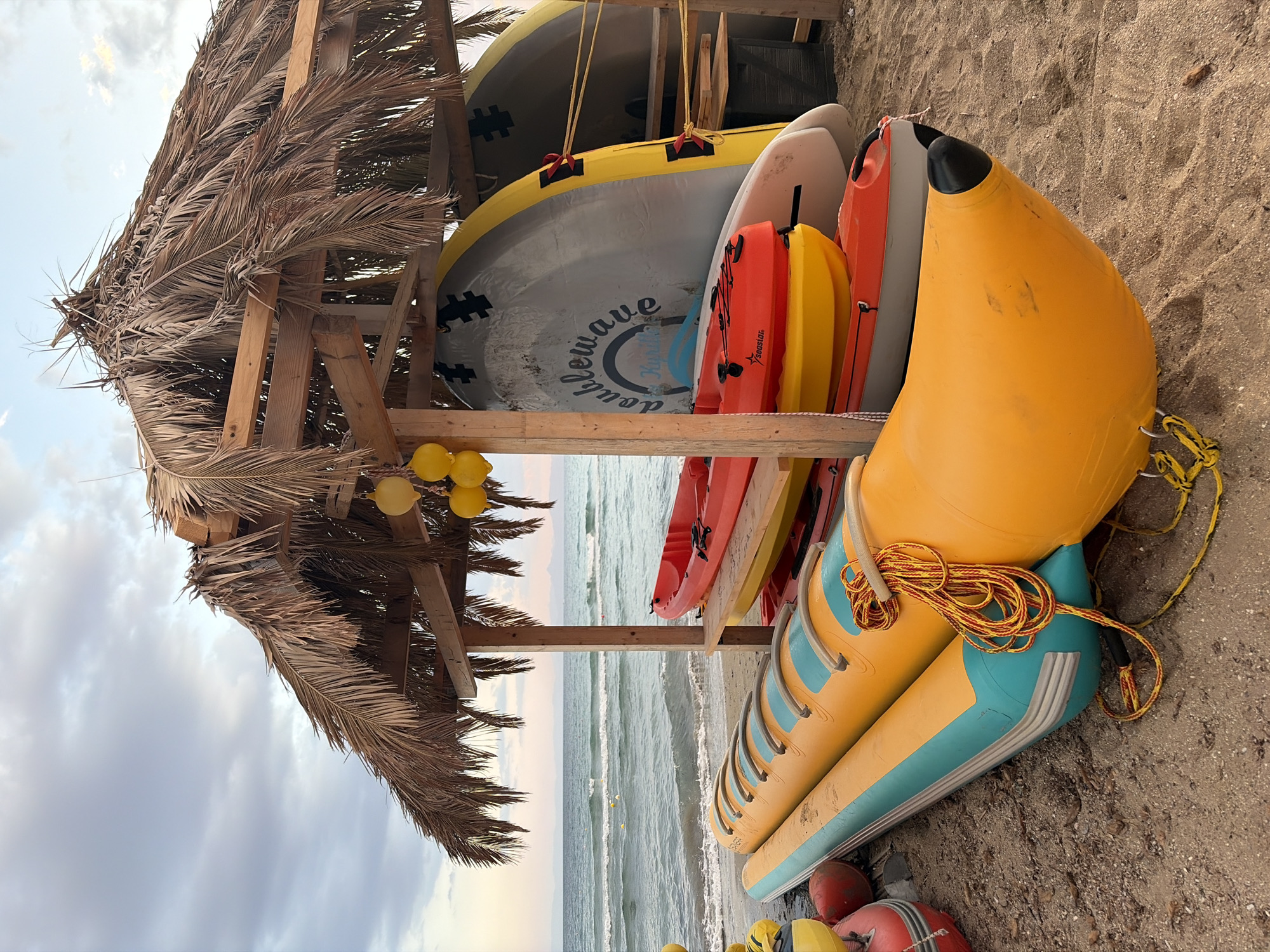 This image, taken in Voula, Greece, captures a serene beach scene featuring water sports equipment neatly stored beside a rustic, thatched hut. The hut is constructed from wooden beams and roofed with dried palm fronds, suggesting a beachside rental or storage facility. Several yellow buoys hang from the hut's wooden frame.
A variety of watercraft are stacked and leaning against the structure and on the sand. These include multiple paddleboards (SUPs), some yellow and grey, with "doublewave" and "Kupala" visible on one's underside, and others in white. Brightly colored kayaks, primarily red and yellow, are also stacked, with the word "Seastart" visible on one of the red kayaks.
In the foreground and to the right, a large, yellow and light blue inflatable water ride, resembling a multi-seater "banana boat," rests on the sand. A coiled yellow and red rope is placed on top of it. Additional red and yellow buoys and another smaller yellow inflatable are partially visible at the bottom of the frame.
The sandy beach extends into the background, where the calm, undulating waves of the sea meet the shore. The sky above is overcast with soft, scattered clouds, indicating a diffused daylight, possibly late morning or afternoon. No people are present, creating a tranquil atmosphere around the organized array of recreational equipment.
