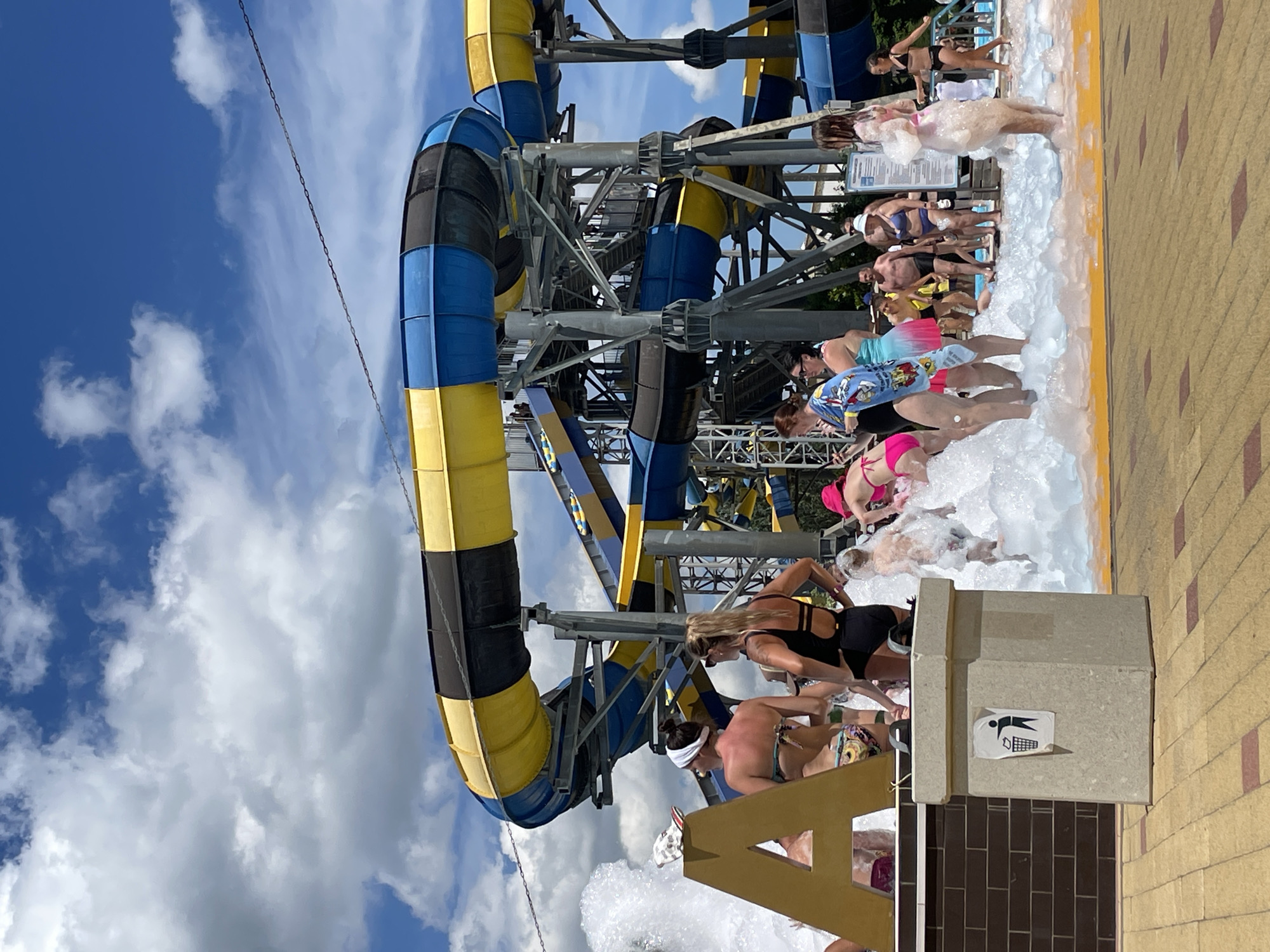 A water park in Anapa, Russia, features a large, multi-level water slide with yellow, blue, and black tubing. The slide is supported by a metal structure. In the foreground, a group of people, primarily women, are gathered in a shallow pool filled with a large amount of white foam. Some are wearing swimsuits and appear to be playing or posing in the foam. A large, golden letter "A" is visible to the left, partially submerged in the foam. A concrete trash receptacle with a recycling symbol is placed in the foreground. The ground is paved with yellow and reddish-brown tiles. The sky above is blue with scattered white clouds.