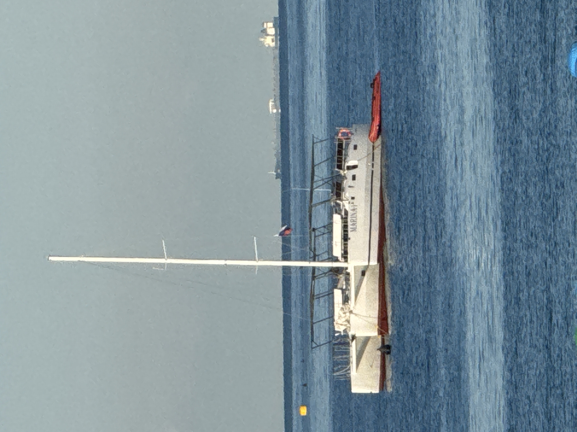 An elevated, medium shot captures a white catamaran with a tall mast sailing on a bright blue body of water under a clear, hazy sky. The catamaran is the primary focus, with its distinctive twin hulls and a white cabin structure. A Russian flag is visible flying from the mast. A bright orange inflatable dinghy is tethered to the side of the catamaran. In the distance, on the horizon, several large cargo ships are visible, suggesting a busy shipping lane. The water is calm, with subtle ripples indicating a gentle breeze. There are no visible people on the catamaran or in the water. The overall impression is one of a peaceful day at sea with maritime activity in the distance.