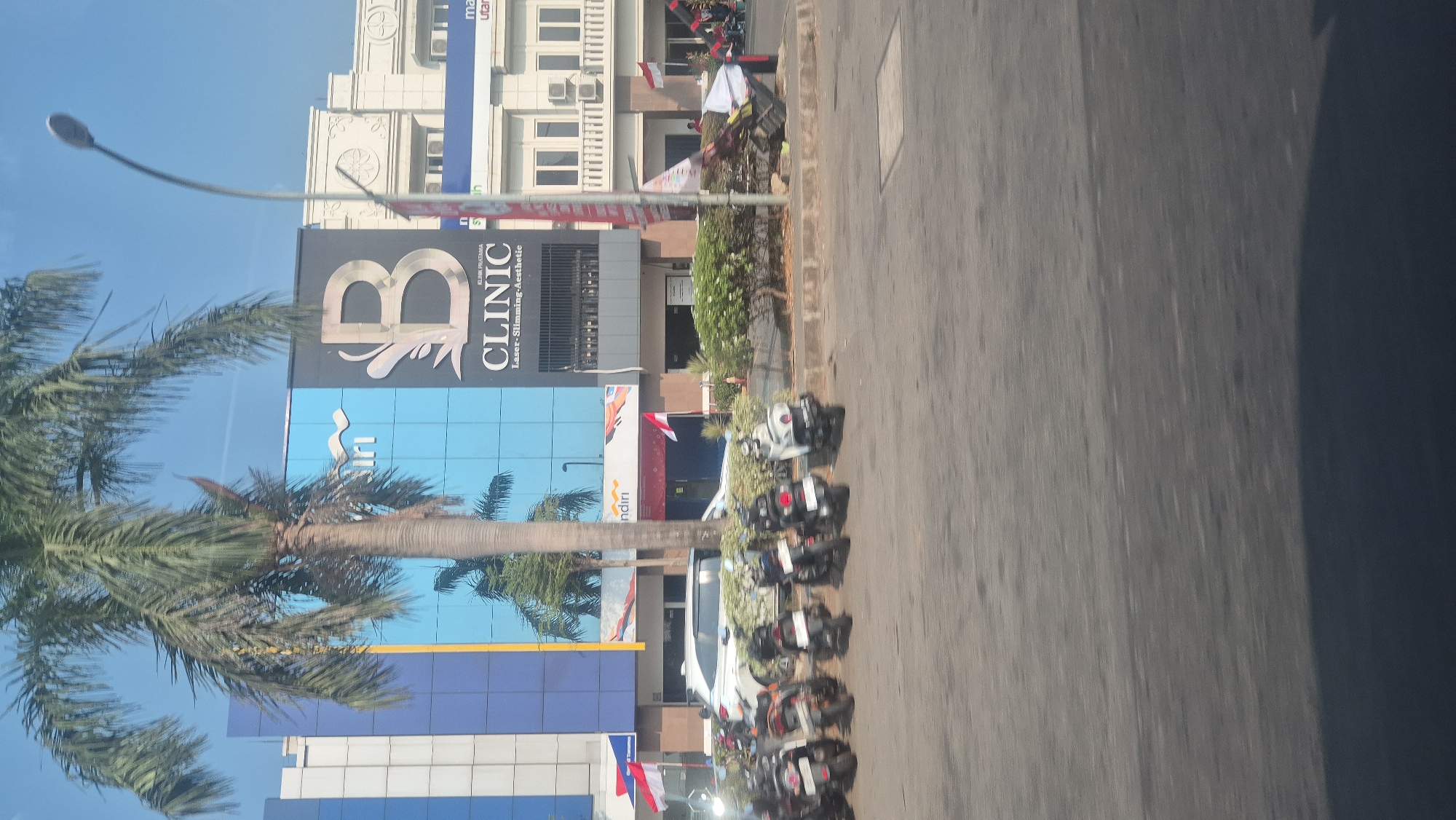 The image shows the front of a modern building complex in Karawang, Indonesia, with a prominent "B CLINIC" sign, specializing in "Laser-Slimming-Aesthetic." The building features a blue glass facade on the left, a dark grey section with the clinic's signage in the center, and a lighter colored building with multiple windows and air conditioning units on the right. In front of the clinic, there are several parked motorcycles and a white SUV. Indonesian flags are visible, including one attached to a pole near the building entrance and smaller flags decorating the building's facade. A tall palm tree stands in front of the blue section of the building. The scene appears to be an outdoor street view with a clear blue sky.