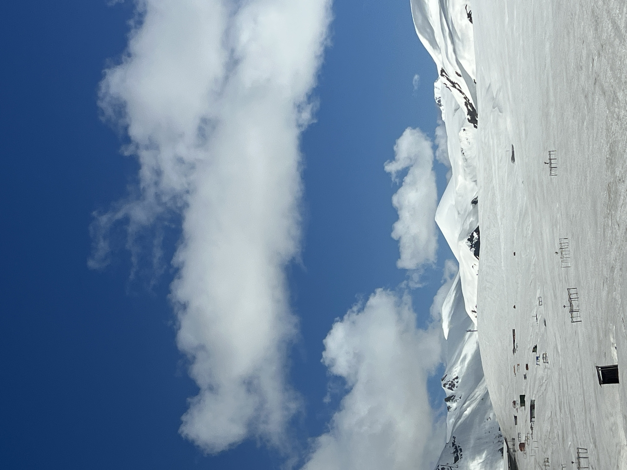 The image depicts a bright, clear day in Ganisi, Georgia, likely in a mountainous region. A crisp blue sky is partially obscured by thick, puffy white clouds. Adjacent to the sky, a snow-covered mountain range stretches into the distance. The foreground shows a snow-covered terrain with sparse infrastructure elements, such as small rectangular structures, possibly ski equipment or buildings. The scene is mostly devoid of human presence.