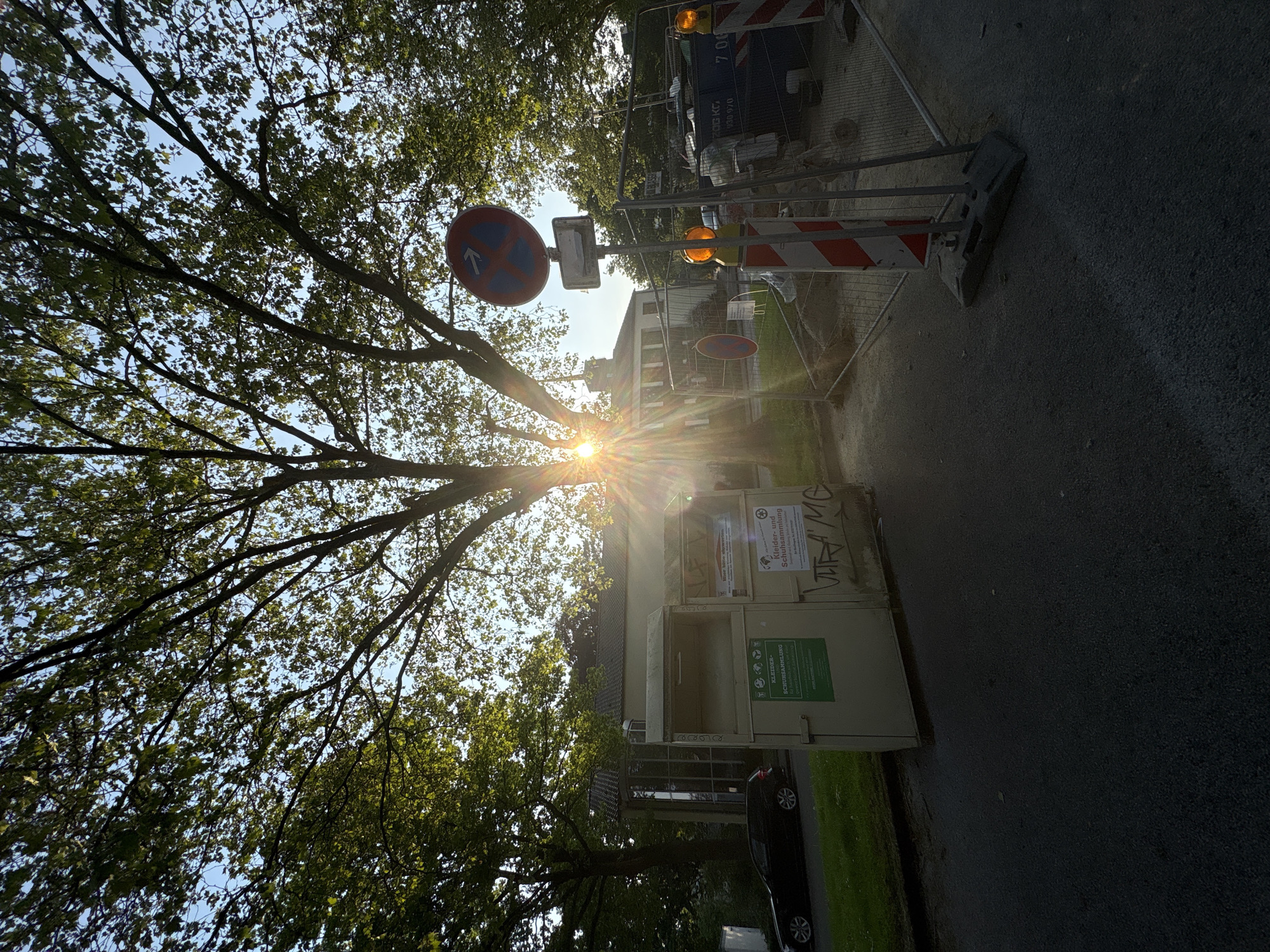 The image depicts an outdoor scene in Mönchengladbach, Germany. A tree with green leaves is visible on the left, partially obscuring the sun, which casts a bright glare. A "no parking" sign is present. To the right of the tree, a clothes donation container stands next to the street. Further along the street, construction barriers and warning lights are erected, possibly indicating road work. A light-colored building is visible in the background. A black car is parked on the grass behind the clothes donation container. The scene is photographed from a low angle, giving a tilted perspective.