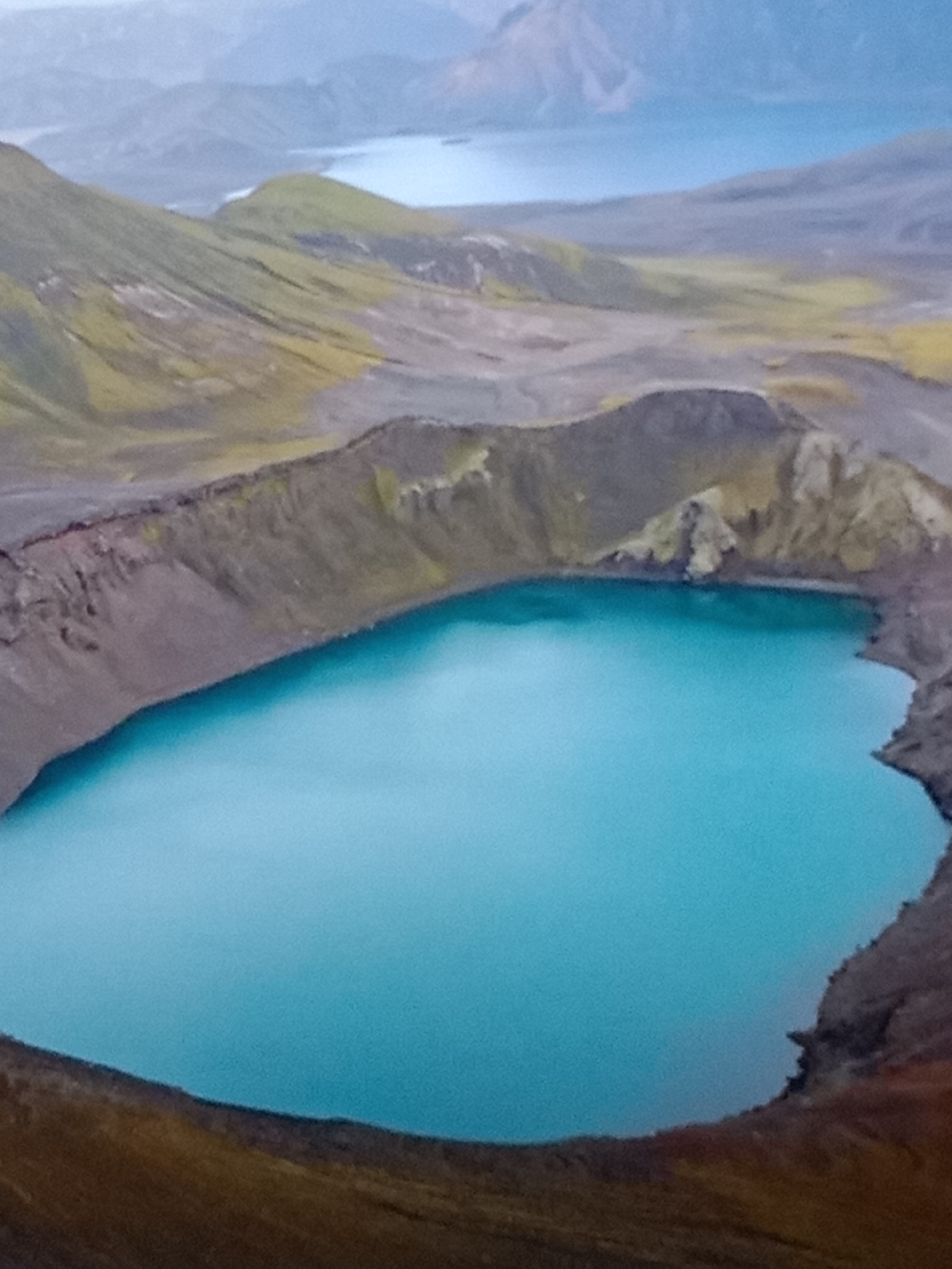 The image shows an aerial view of a turquoise lake nestled within a caldera-like formation. The surrounding landscape is characterized by steep, eroded slopes covered in patches of green and yellow vegetation and rocky, barren terrain. In the background, a hazy, mountainous landscape with another body of water is visible. The scene is devoid of any human presence or identifiable structures. It is not possible to determine the location in the city of Kielce, Poland, as the image depicts a natural geological formation.