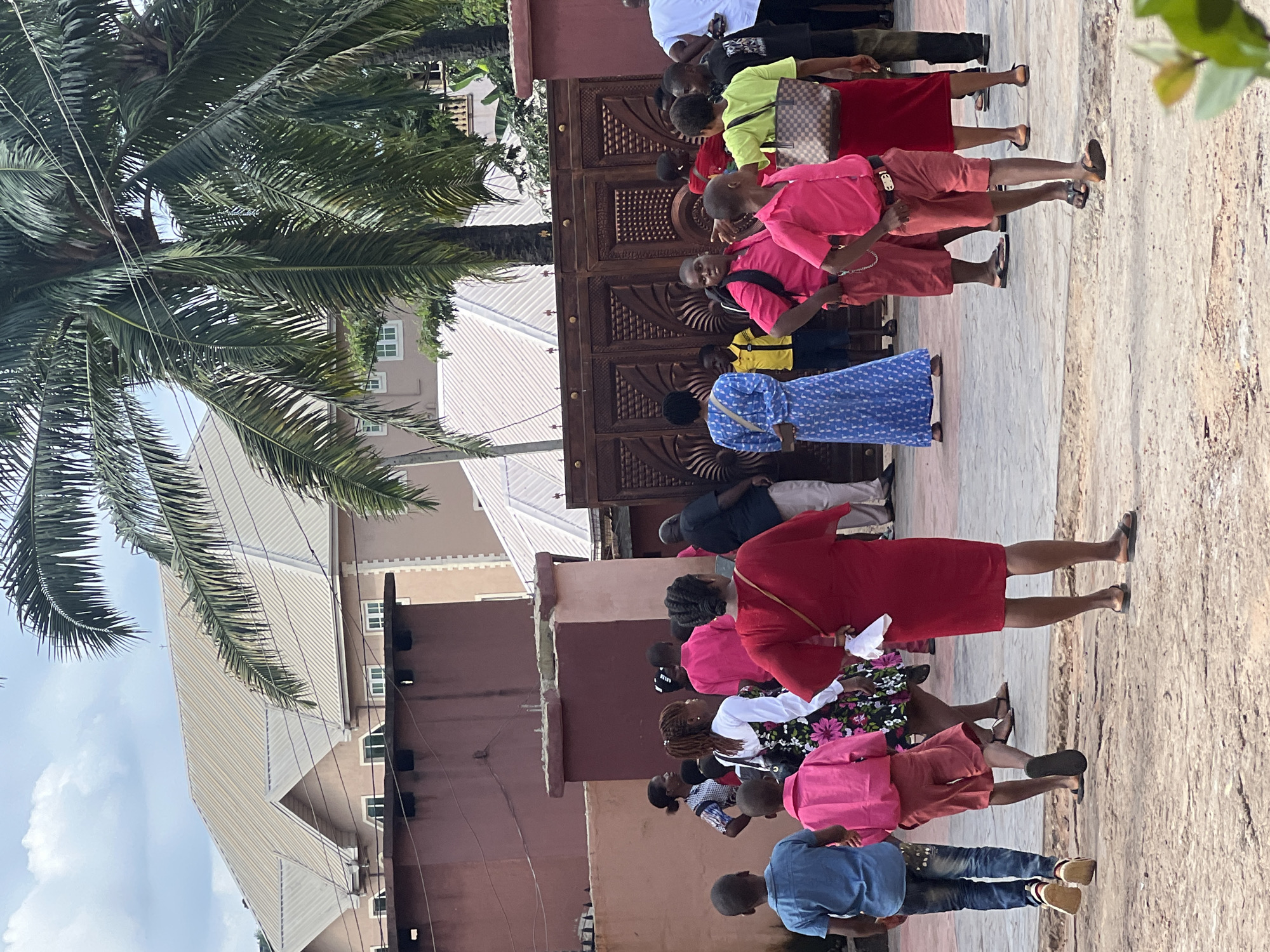 The image depicts an outdoor scene in Nnewi, Nigeria, likely in front of a building with a large, ornate wooden gate. A group of people, including children and adults, are gathered on a paved area. Several individuals are wearing bright, colorful clothing, such as pink shirts and red dresses. Palm trees are visible in the background, and there are multi-story buildings with light-colored roofs. The ground appears to be a rough, sandy texture. The overall impression is of a casual gathering or a community event.