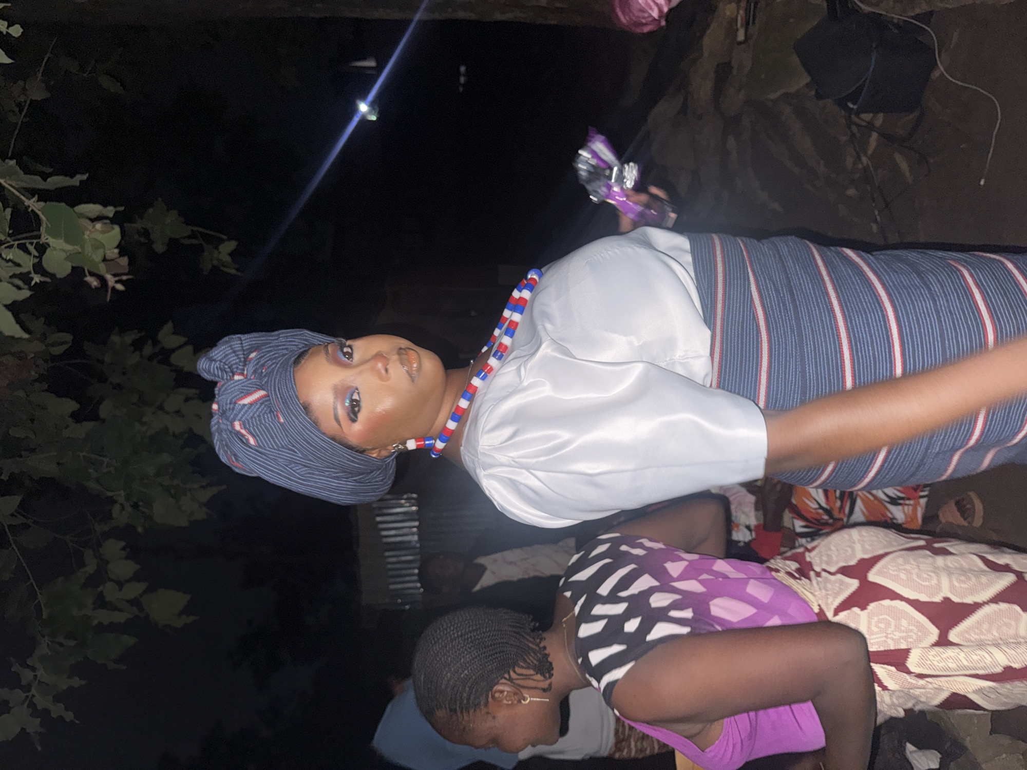 A woman is captured in a medium shot, standing outdoors at night in Gura Top, Nigeria. She is adorned in traditional attire, featuring a blue and white striped headwrap, a white puffed-sleeve top, and a blue, white, and red striped skirt. She wears a multi-colored beaded necklace and earrings that complement her outfit. Her makeup is noticeable with bold blue eyeshadow. She holds a purple object in her right hand. To her left, another woman is visible, wearing a pink tank top and a patterned wrap skirt. The background is dark, suggesting an evening or nighttime setting, with trees and foliage in the upper left. Other individuals are partially visible in the background, and a black bag is on the ground to the right.