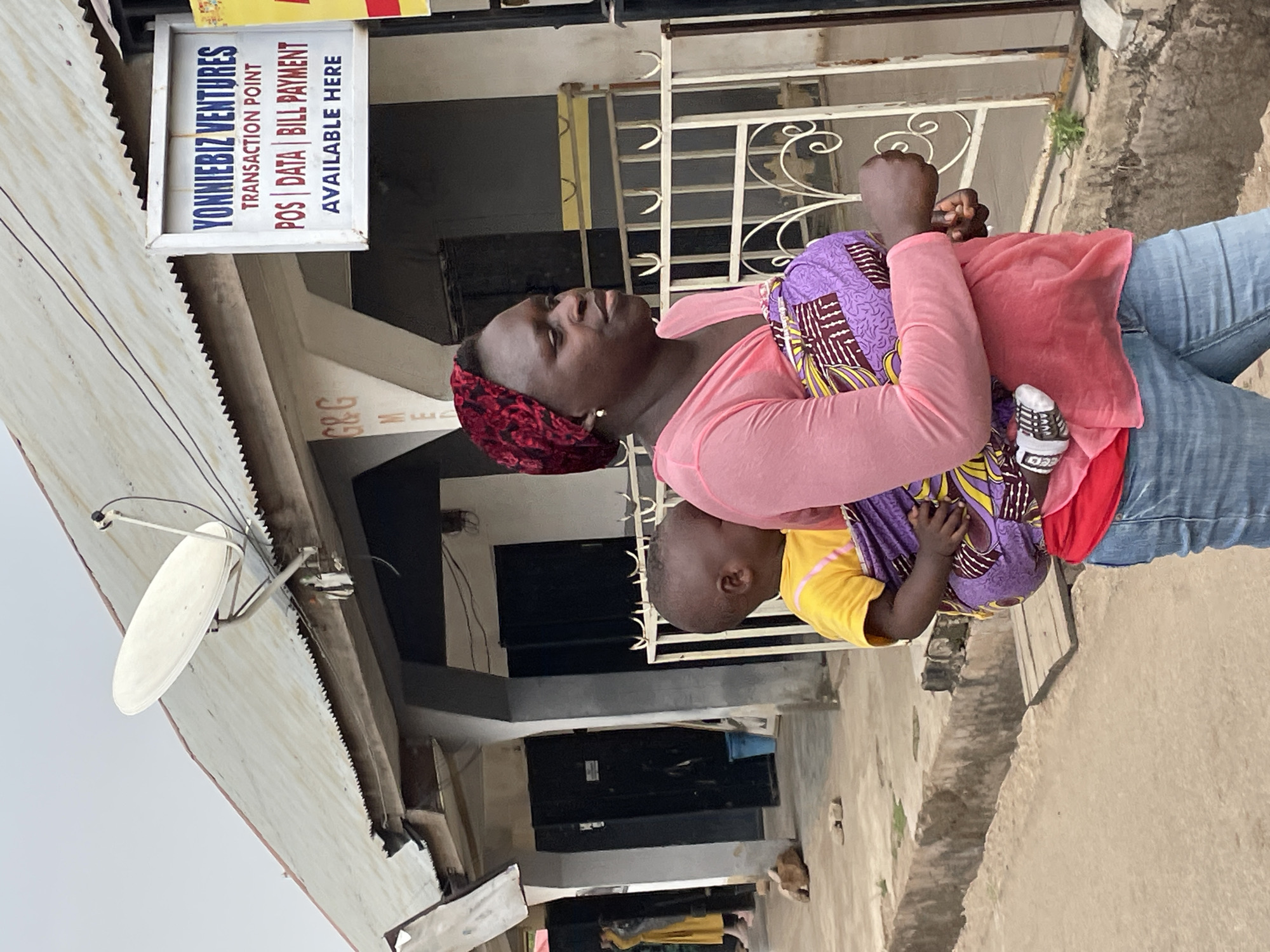 A woman is carrying a young child on her back in front of a building with a sign reading "YONNIEBIZ VENTURES" in Jos, Nigeria. The woman is wearing a pink long-sleeved shirt and blue jeans, with a red headscarf. The child is wearing a yellow shirt and is secured to her back with a purple and yellow patterned fabric. The sign indicates that YONNIEBIZ VENTURES offers transaction point services, including POS, data, and bill payment. A satellite dish is visible on the roof of the building.