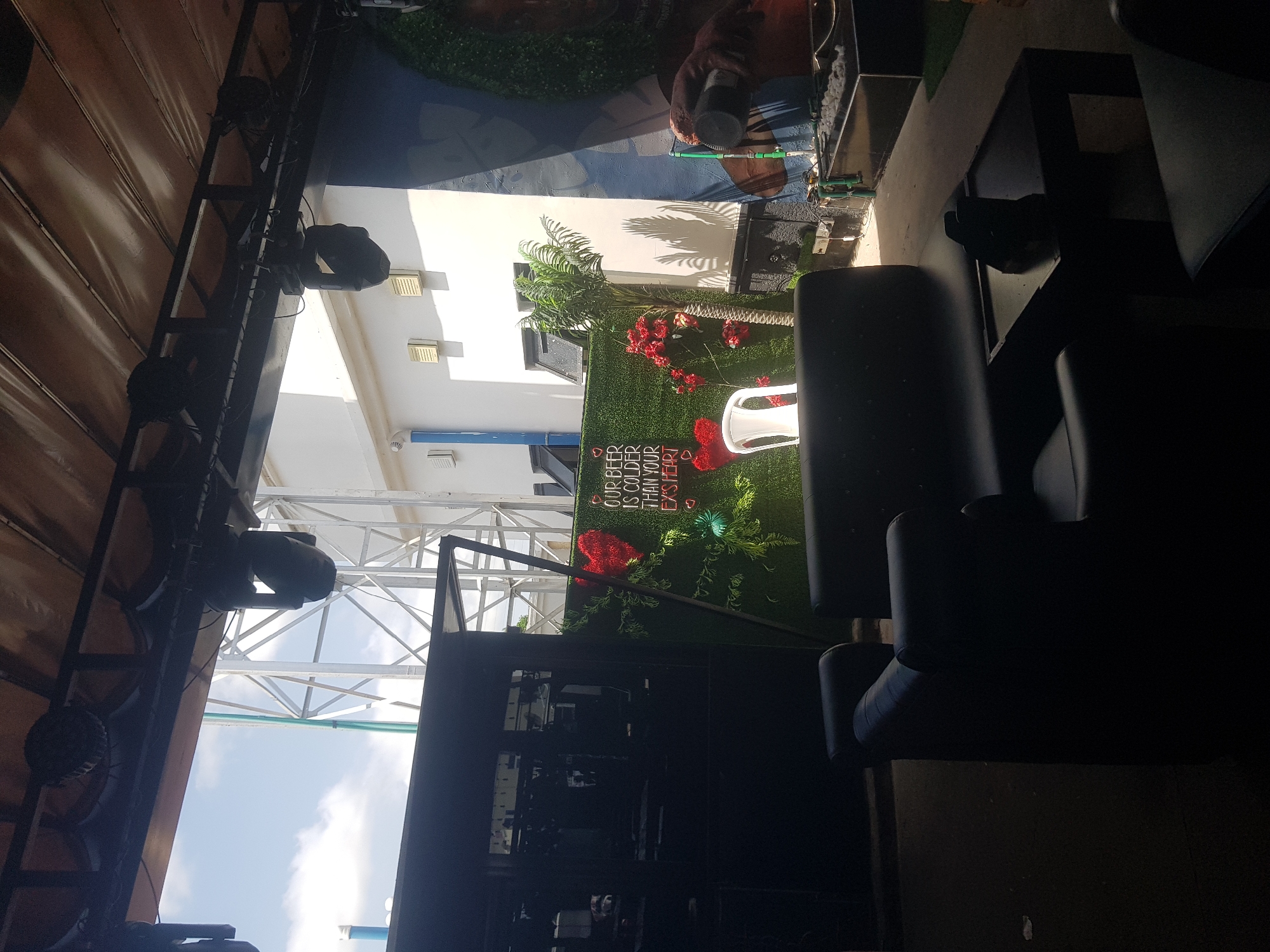 The image depicts an outdoor seating area, likely a bar or lounge, with a backdrop of artificial greenery and a neon sign. The neon sign reads: "OUR BEER IS COLDER THAN YOUR EX'S HEART." In the foreground, there are dark leather sofas and a small black table. A white plastic chair is positioned in front of the green wall. Overhead, lighting equipment is suspended from a canopy. Beyond the seating area, a white building and a large metal structure are visible against a partly cloudy sky. The setting appears to be in Howayi, Nigeria, suggested by the context provided, though this is not visually evident from the image itself.