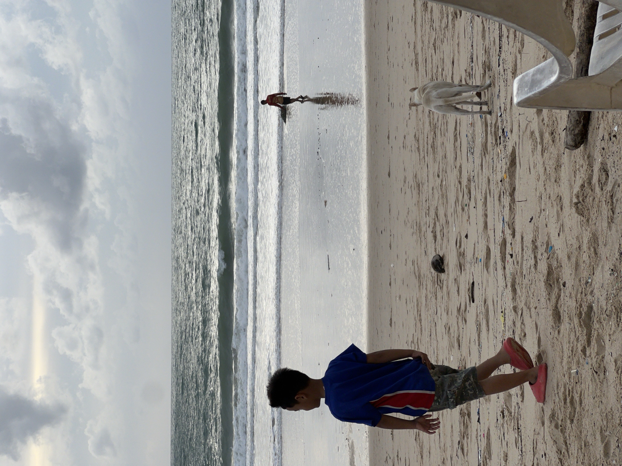 A boy in a blue and red shirt and camouflage shorts walks on a wet sandy beach. He is facing away from the camera. A white dog stands on the sand in the middle ground to the right. In the background, a person carrying a surfboard walks into the ocean. The sea is choppy with white waves under a cloudy sky. The image appears to be taken at Surin Beach in Choeng Thale, Thailand.