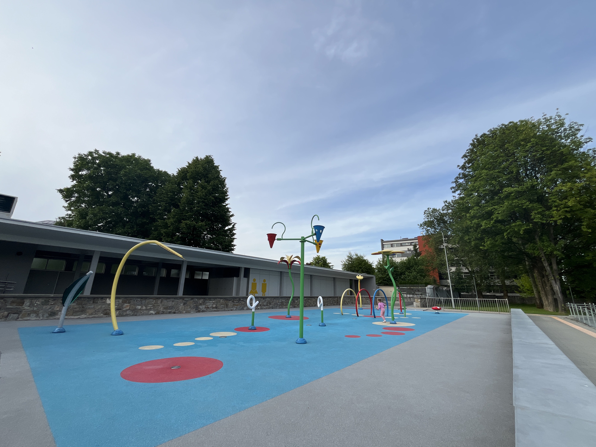 The image displays a colorful and modern splash pad, likely located in a public park or recreational area. The ground features a blue rubberized surface with scattered red and yellow circular patterns. Numerous water play structures are visible, including a prominent central green pole with multiple colorful buckets and nozzles, a curved yellow arch, smaller arched tunnels in red, yellow, and green, and white loop-shaped sprayers. Tall, stylized flower-like structures also contribute to the playful design. No water is visibly flowing from the features at the moment the photo was taken.
A single young child, appearing to be a girl in a pink outfit, is visible on the right side of the splash pad, seemingly running or walking among the play structures.
In the background, a modern, low-rise building with a grey facade, large windows, and a stone-clad base runs along the length of the splash pad. Large white graphic icons, representing generic male and female figures, are visible on the building's wall, indicating restrooms or changing facilities. Lush green trees flank the scene on both sides and extend into the background. A multi-story residential building is partially visible beyond the trees on the right. A grey concrete pathway or low wall borders the right side of the splash pad.
The sky is overcast with some visible cloud formations, suggesting a daytime scene under a calm, perhaps slightly cloudy, weather condition. The overall atmosphere appears inviting and designed for children's outdoor play.