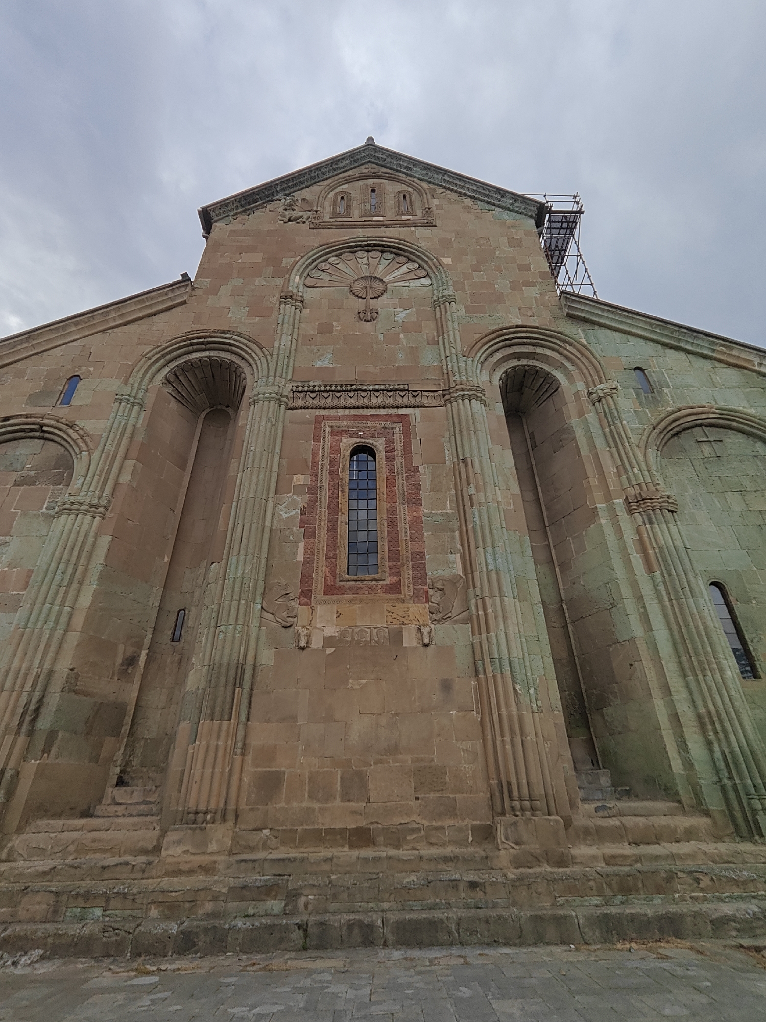 A low-angle, upward-looking shot captures the imposing stone facade of an ancient building, likely a church or monastery, under an overcast sky. The building, constructed from large, rough-hewn blocks, exhibits varying shades of brown, tan, and a distinctive greenish patina on some surfaces, suggesting age and weathering.
The central part of the facade features an elongated, rectangular window with a grid pattern, set within a recessed panel decorated with red and yellow geometric and floral carvings. Above this, a large circular rosette-like carving is visible, flanked by smaller arched windows further up towards the gable.
On either side of the central window, deep arched recesses are present, framed by fluted pilasters that extend downwards towards the base of the structure. Some of these recesses contain smaller, narrow windows. The architectural style appears to be medieval or early modern.
Scaffolding is visible on the upper right side of the roofline, indicating ongoing maintenance or restoration work. The ground level shows paving stones, and the base of the building is made of larger, rougher stone blocks, forming a foundation. No people or discernible text are visible in the image.