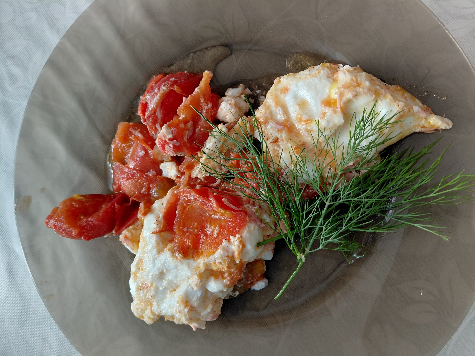 A close-up overhead shot shows a dish of cooked eggs and tomatoes, garnished with a sprig of dill, served on a translucent brown plate. The plate rests on a white, textured tablecloth. The dish appears to be a simple breakfast or light meal. The composition focuses on the food, with the background blurred. There is no visible indication of location in Tashkazgan, Russia.