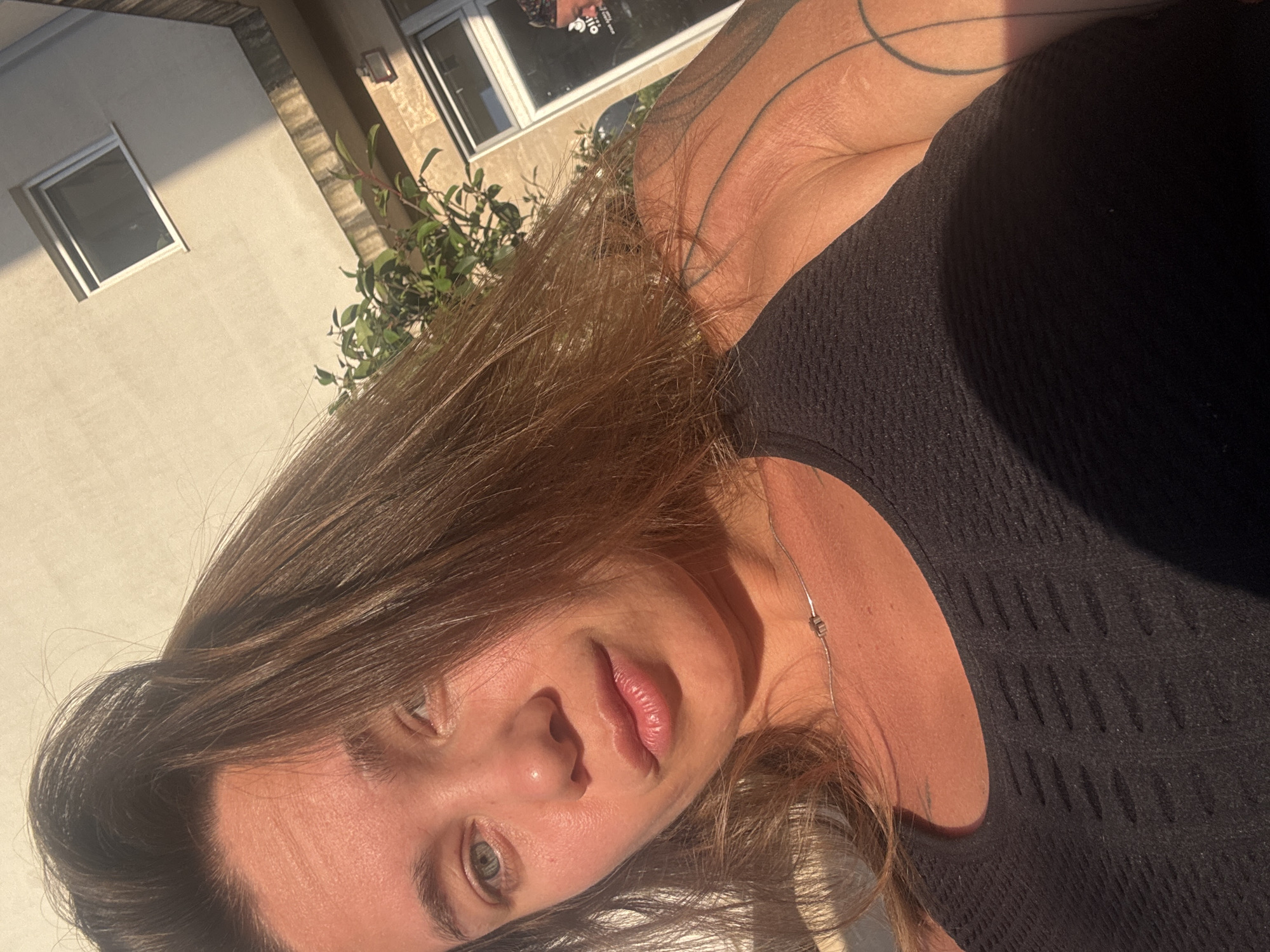 The image presents a close-up, diagonally oriented portrait of a person, likely female, captured in natural daylight. The subject has medium-brown hair that frames her face and light-colored eyes, possibly blue or green. Her lips are full, and her complexion appears tanned. She wears a dark (likely black) textured top or dress, and a thin silver necklace with a small charm is visible around her neck. Faint tattoo outlines can be seen on her upper arm/shoulder. Her expression is neutral and relaxed, suggesting a moment of rest or a casual pose for the camera.
The background elements suggest an outdoor setting in Budva, Montenegro. Visible behind the subject are parts of a light-colored building with modern white-framed windows. Some green foliage also peeks into the frame, adding to the outdoor ambiance. The strong, warm sunlight indicates it is daytime, possibly morning or late afternoon, creating distinct highlights and shadows across the subject's face and surrounding details. In the reflection of one of the windows, some blurry and indistinct text, possibly from a sign or poster, can be vaguely discerned. No clear, readable text is present. The overall scene conveys a casual and sunlit portrait.