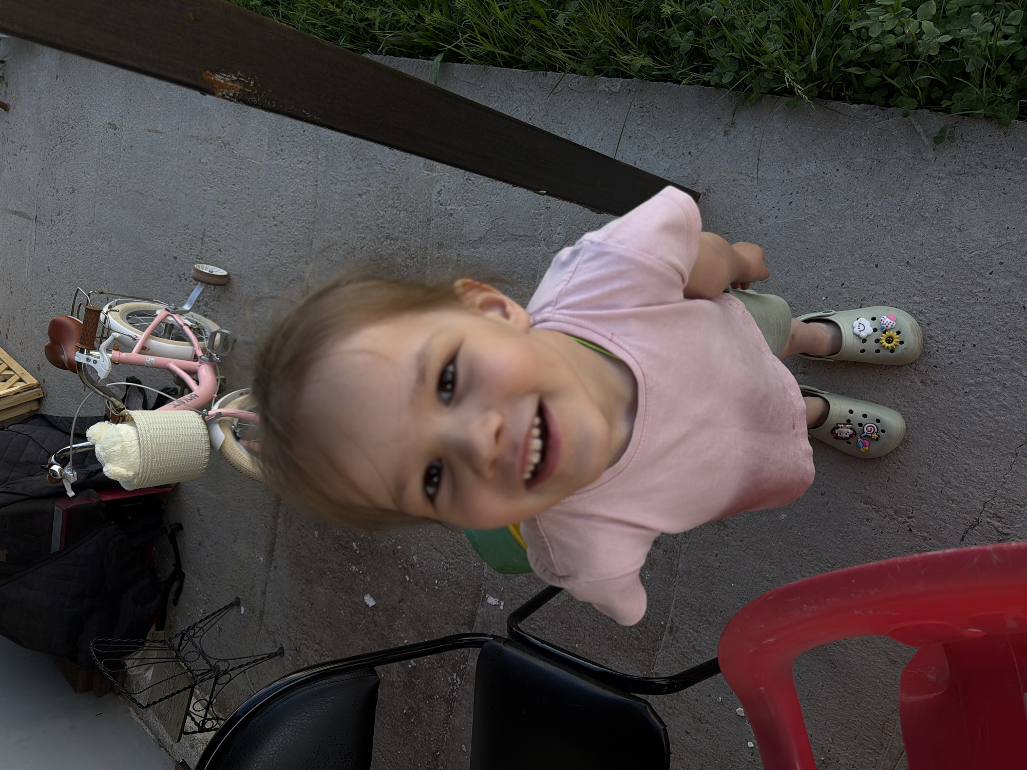 The image depicts a young child in Chaikovsky, Russia, smiling and looking up at the camera. The child is wearing a pink shirt, light green shorts, and decorated light green Crocs-style shoes. The child is standing on a concrete surface. In the background, there is a fence, grass, a small pink bicycle with a basket, and a black bag. A red plastic chair and a black metal chair are also visible.