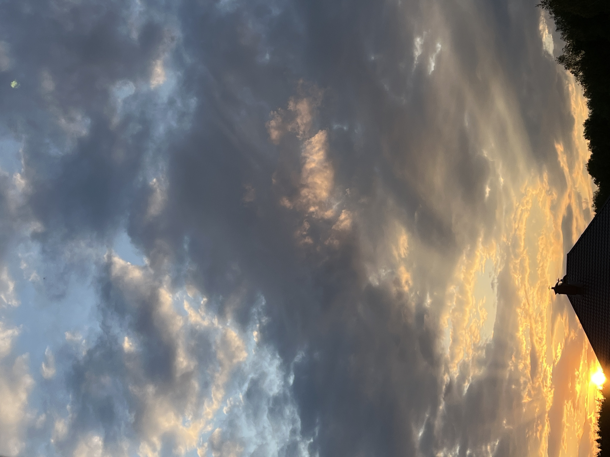 The image depicts a sunset sky with a mix of dark, heavy clouds and areas where the sun is breaking through, casting a golden light. Some trees are visible at the top right, along with a building's roofline featuring a weathervane. The image appears to have been taken in an urban environment. The exact location within the city of Unknown, Unknown is unidentifiable based solely on this image.