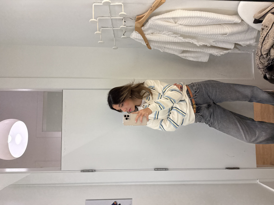 A woman stands in a mirror, holding a smartphone and taking a picture of herself. She wears a striped sweater and gray jeans with a brown belt. Above her, on the right side, a coat and a hanger are visible. In the upper left corner, there's a white coat rack. To the left, a portion of a room with a white, spherical lamp is visible in the mirror's reflection. The background consists of white walls and a doorway.