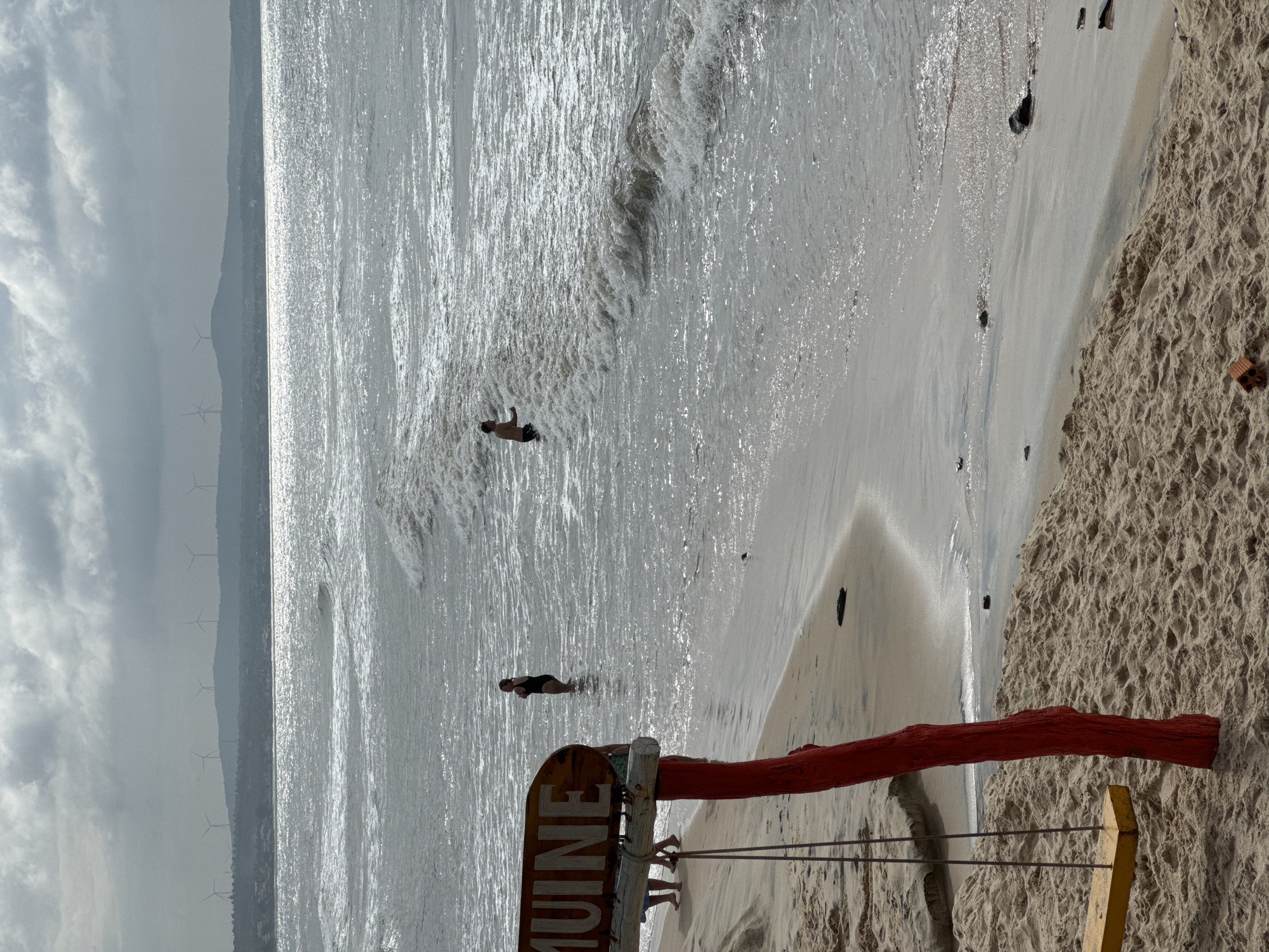 The image depicts a coastal scene in Mũi Né, Vietnam, likely during overcast weather, indicated by the cloudy sky and diffused sunlight. In the foreground, a yellow wooden swing hangs from a weathered wooden post painted red, set against a sandy beach. To the left, a wooden sign with the partially visible word "MUINE" is mounted on the red post.
The middle ground features the ocean with moderately sized waves rolling towards the shore. Two individuals are visible in the water. One, wearing a black swimsuit, stands waist-deep in the water on the left side of the frame. Further out, a person with no shirt on is partially submerged, facing away from the camera and towards the waves. Several other figures, possibly a group of people, are visible near the shoreline on the left, their lower bodies in the sand.
The background shows a hazy horizon line with landmasses and what appear to be wind turbines silhouetted against the sky. The overall atmosphere is subdued, with muted colors and a slightly muted light quality. The beach appears sandy, with some debris visible along the waterline.