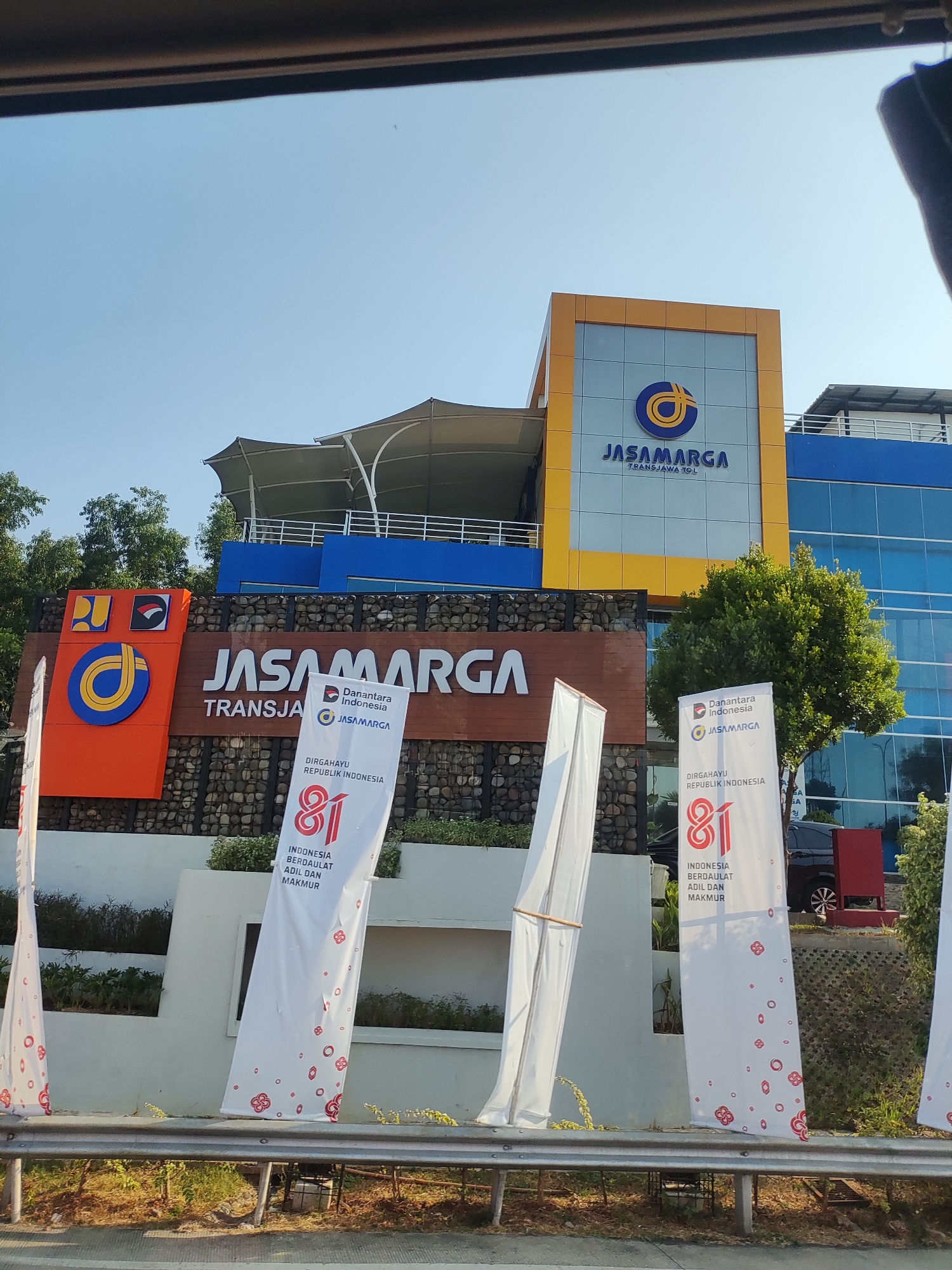 The image shows the Jasa Marga Transjawa Tol office building in Karawang, Indonesia, on a clear day. The building features a prominent yellow and blue facade with the Jasa Marga logo. Several large white banners with red lettering and graphics are displayed in front of the building. One banner prominently features the number "81" along with text related to Indonesian Independence Day. The building is surrounded by trees and landscaping. A guardrail is visible in the foreground, suggesting the photo was taken from a vehicle.