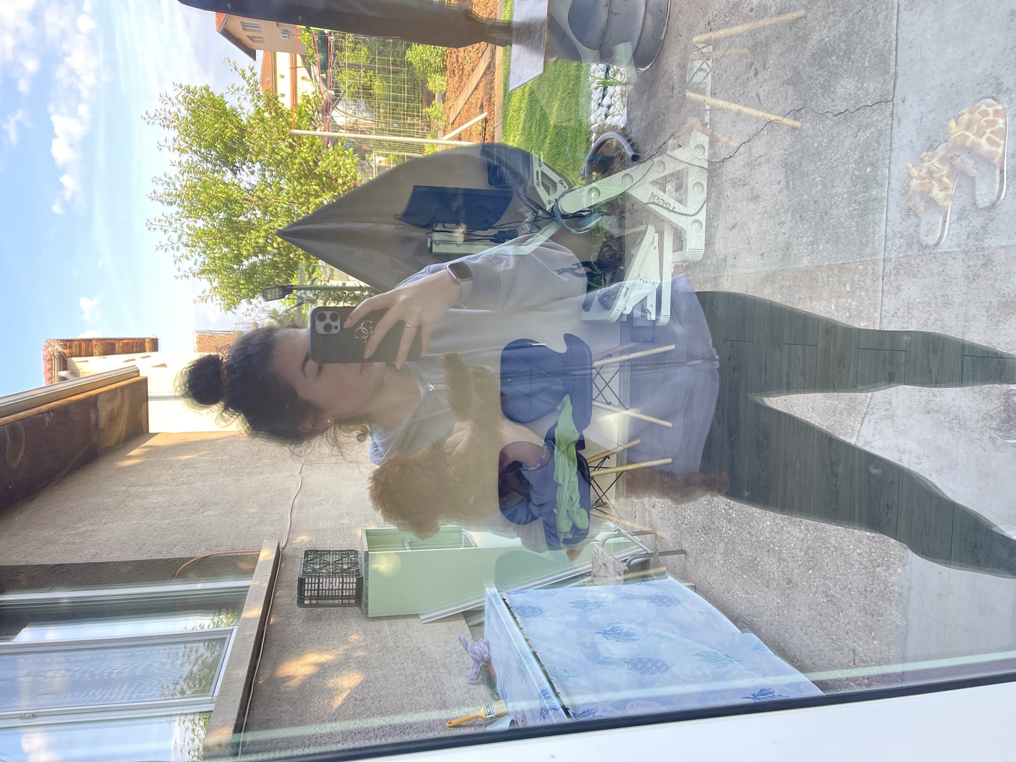 A woman with dark hair styled in a bun takes a photo with her cell phone, reflected in a window. The reflection shows she is wearing leggings and a hoodie, standing in an outdoor setting. Visible through the window are objects including a lawn mower, a clothes drying rack with clothes, a painted dresser, and stuffed animal slippers on a concrete surface. The environment includes buildings, trees, and a blue sky with clouds. The location appears to be Sofia, Bulgaria, based on the architecture and environment visible outside the window.
