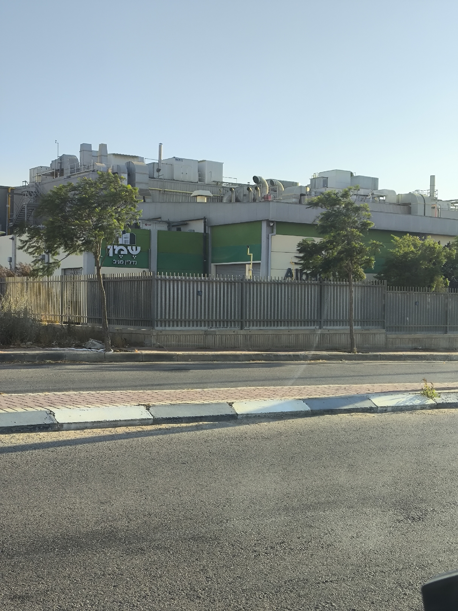 The image captures a view of the "Tnuva" dairy processing plant located in Dimona, Israel. The plant is a large industrial building with a flat roof covered in numerous ventilation units and machinery. The main structure is painted in sections of white and green. In front of the building is a metal fence and a single, slender tree stands in the foreground. The surrounding area appears to be an urban or industrial zone, with a paved road and sidewalk in front of the fence. The sky is clear and blue, suggesting a sunny day.