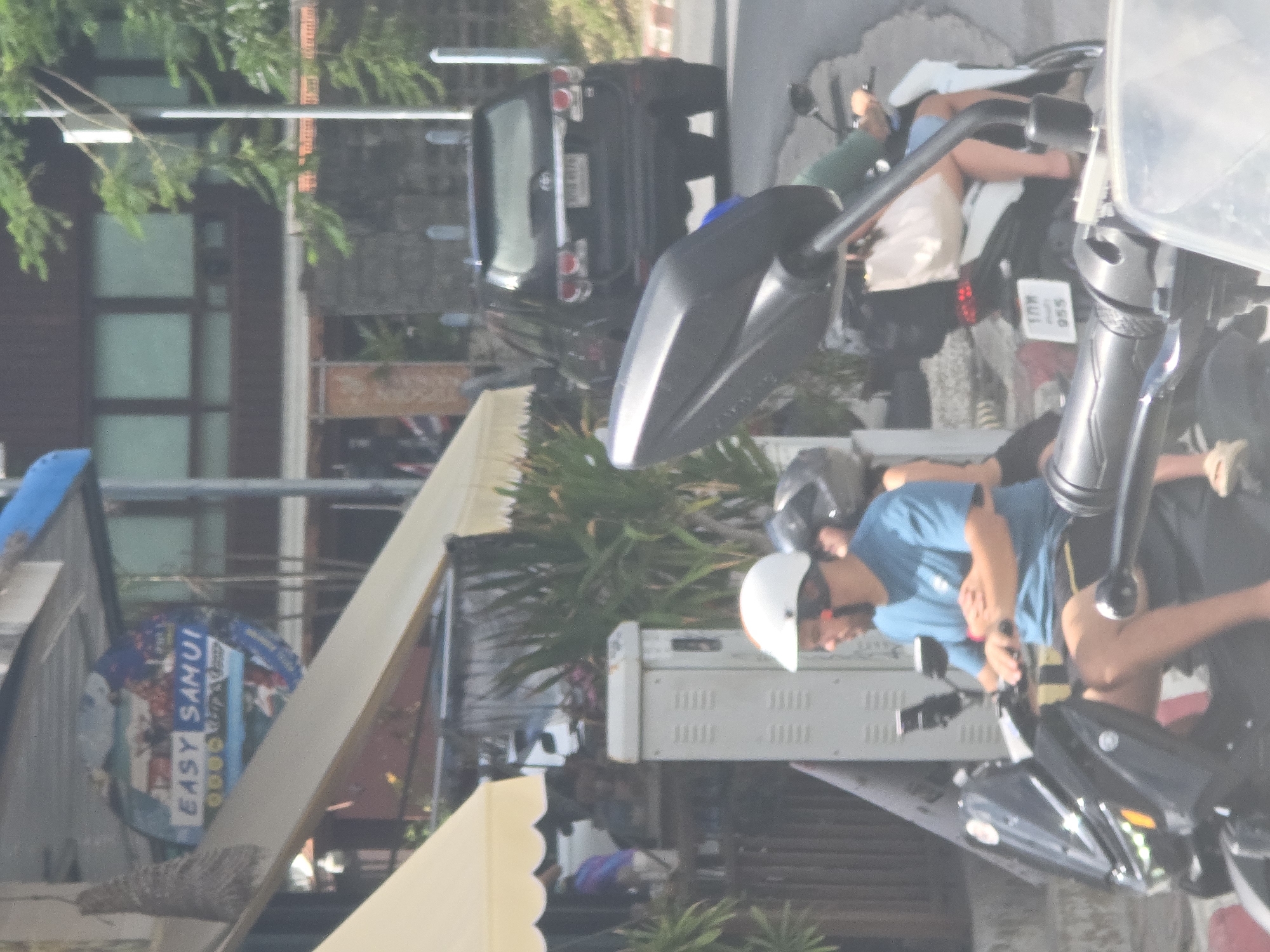 In a city scene, a man wearing a white helmet and a blue t-shirt rides a black scooter. He is looking forward, with his hands on the handlebars. Behind him, another scooter is visible with a rider wearing a white helmet and a green shirt. A black SUV is parked on the side of the road in the background, with a license plate that is partially visible. The environment appears to be a street with buildings and trees. A sign that says "EASY SAMUI" is visible on a building in the distance.