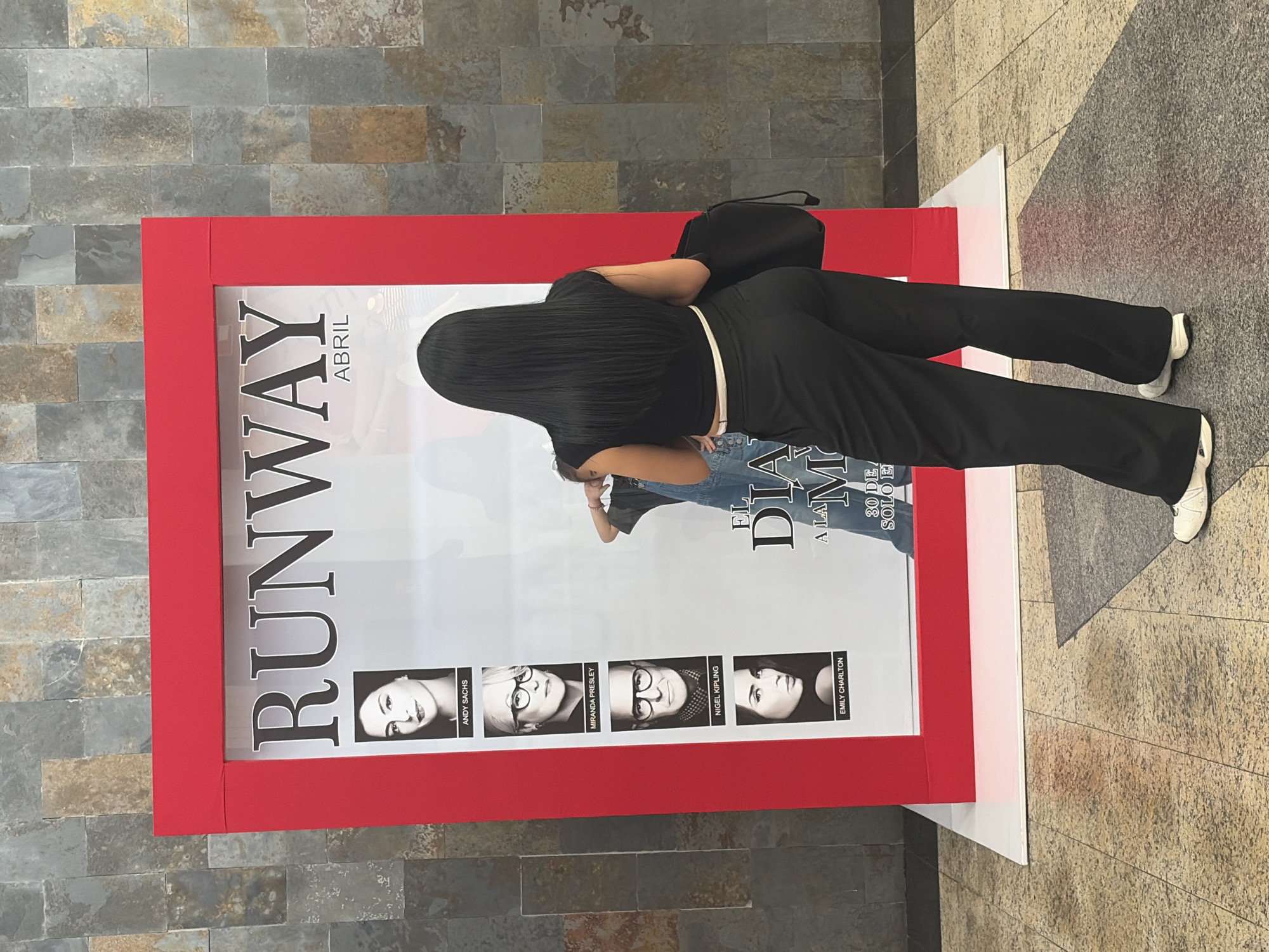 A woman in black pants and a black shirt is posing next to a red-framed poster for a "Runway" event in April, featuring photos of Andy Sachs, Miranda Presley, Nigel Kipling, and Emily Charlton. A second person is visible behind the woman taking the photo. The poster is located indoors on a tile floor, and the background includes a wall of gray stone tiles. The city is Ciudad Guayana, Venezuela.