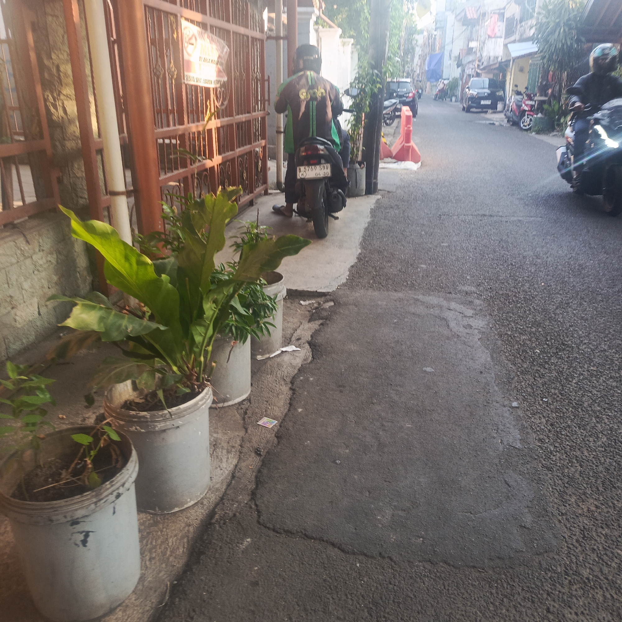 The image depicts a narrow urban street scene in South Jakarta, Indonesia, bathed in the warm light of what appears to be early morning or late afternoon. In the foreground, several potted plants are situated along a narrow sidewalk. The plants in the largest pots are lush and green, with broad leaves.
The street itself is paved with asphalt and shows signs of wear. A person wearing a black and green jacket with the "Grab" logo, a ride-hailing service, is seated on a black motorcycle. The license plate of the motorcycle is visible and reads "B 5769 SBV". The rider is facing away from the camera, suggesting they are either waiting or about to depart.
Further down the street, other vehicles, including cars and motorcycles, are parked or in motion. Red and white traffic barriers are placed along the side of the road, indicating some form of demarcation or ongoing work. Buildings line the street, exhibiting typical Indonesian urban architecture. The overall atmosphere is that of a bustling residential or commercial area in a dense city. A sign with a "no smoking" symbol is visible on a fence in the background, suggesting a private property boundary.