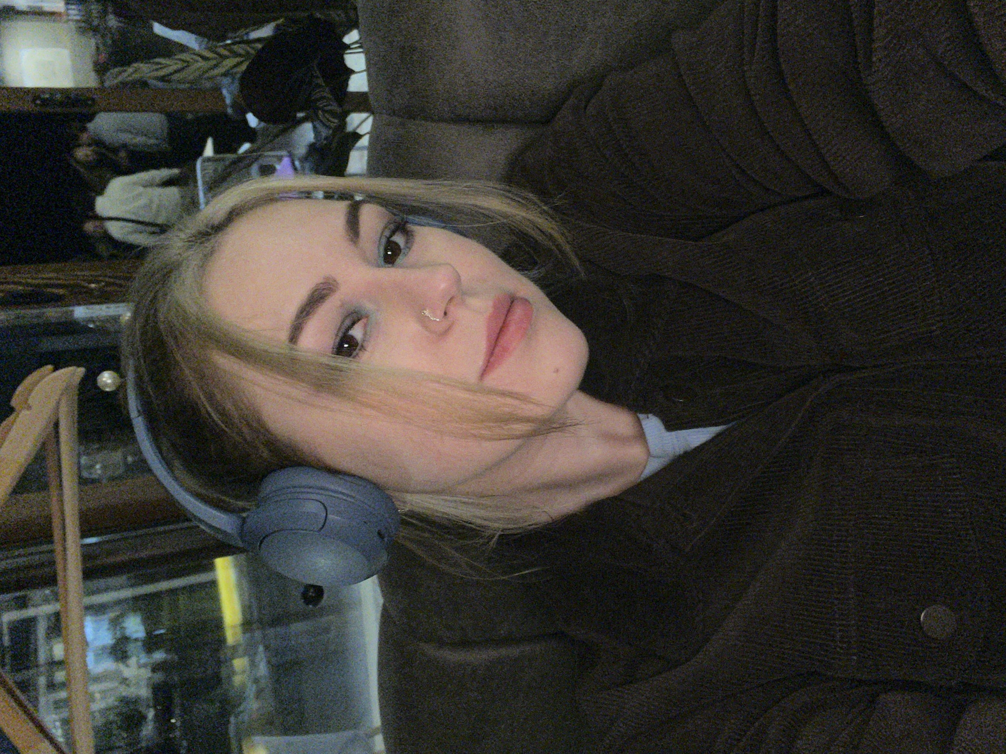 A woman with blonde hair and headphones is reclining on a brown upholstered couch. She has a nose piercing. She's wearing a brown corduroy jacket over a blue shirt. In the background, there is a reflective surface showing a blurred scene with figures, likely a bar or restaurant setting. The location mentioned, Boreti, Montenegro, is not visually discernible from the image.