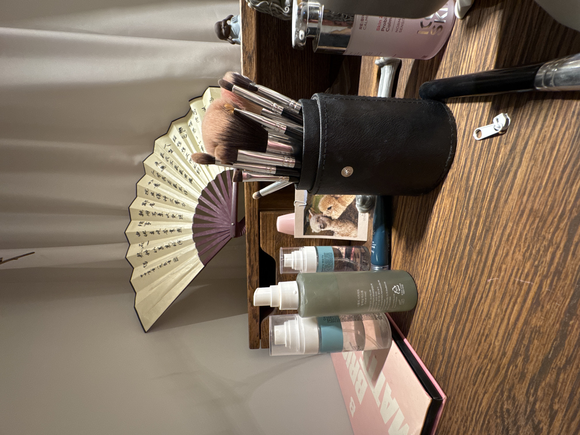 The image shows a close-up view of a wooden desk with various cosmetic items. In the foreground, a black cylindrical container holds a collection of makeup brushes with dark handles and fluffy tips. To the left of the brushes, there are three clear plastic bottles with spray tops, two of which contain liquids, and one is empty. A pink eyeshadow palette is positioned to the far left. Behind the brushes, a portion of an open fan with intricate black calligraphy is visible, leaning against a cream-colored curtain. To the right of the brushes, a light pink bottle with white text is partially visible, and a small picture frame featuring two llamas is placed further back. A dark blue tube and a white zipper pull are on the desk near the brushes. The overall lighting suggests an indoor setting, possibly a dressing table or vanity. The location is stated to be in Бараўлянскі сельскі Савет, Belarus.
