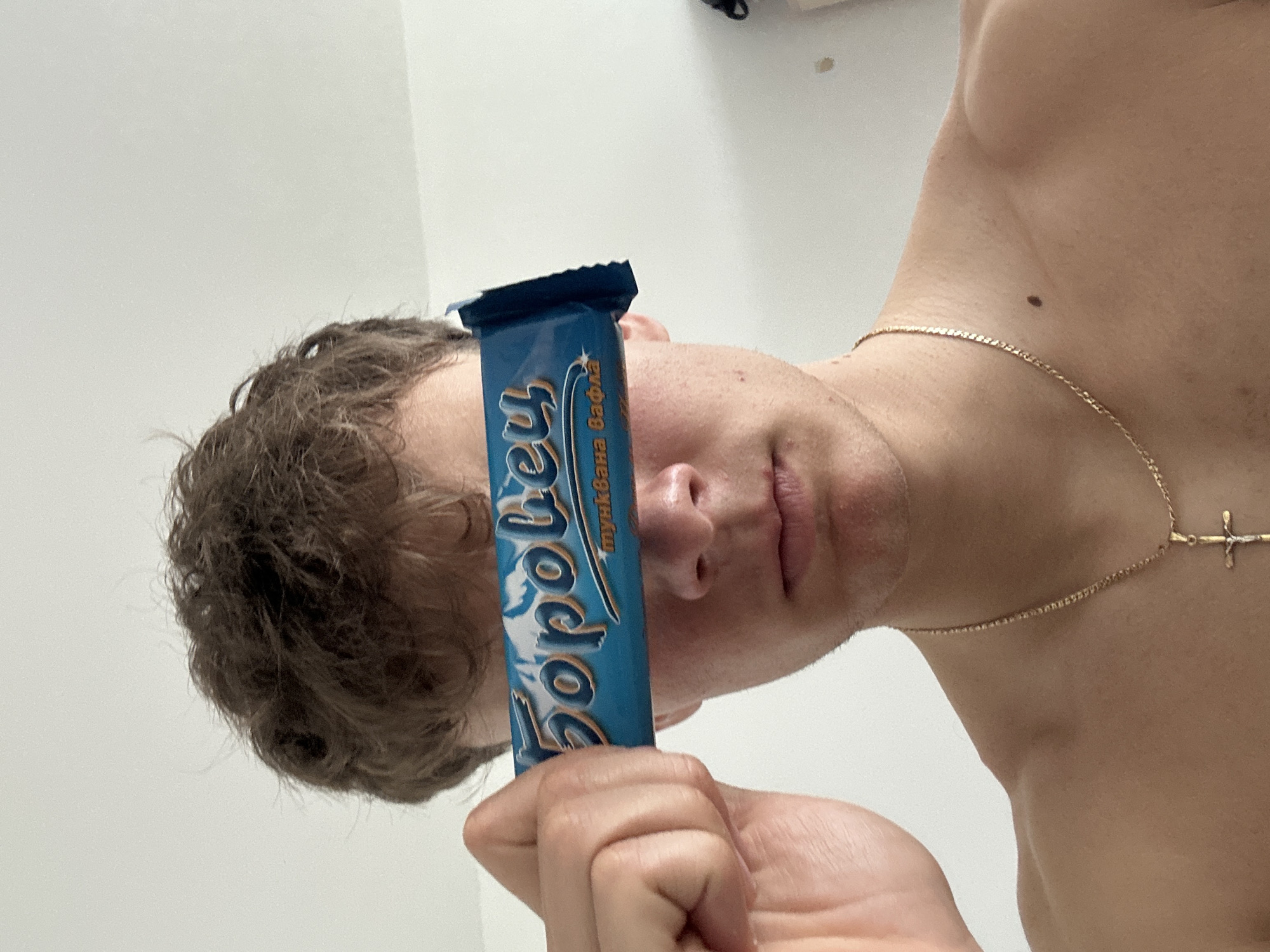 A man is holding a blue candy bar with the word "Topokey" on it in front of his face. He has brown hair, a fair complexion, and is wearing a gold necklace with a cross pendant. The background is a white wall. Location: Unknown, Unknown.