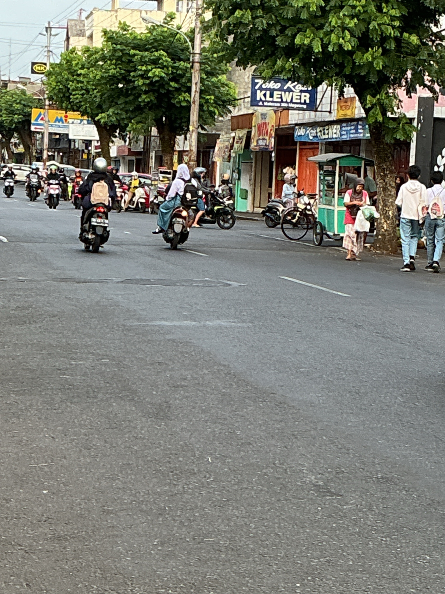 The image depicts a street scene in Magelang, Indonesia, likely on a main road given the traffic and commercial establishments. The road is paved and appears to be busy with several motorbikes in motion. In the foreground, the asphalt road surface is visible, with a white line marking a lane.
On the right side of the road, there are buildings and shops. One prominent sign reads "Toko Kain KLEWER." A small green food stall is also present, with a woman in a red top and patterned skirt standing near it, holding a plastic bag. Further down the street, three young people, two males and one female, are walking away from the camera. The males are wearing white hoodies and jeans, while the female is wearing a white shirt and jeans.
On the left side of the road, more motorbikes are seen. A person wearing a white helmet and a beige backpack is riding a motorbike. There are also shops and signs visible in the background, including a yellow sign with "HK" on it and a blue sign with a red and white symbol. Lush green trees line the street, providing shade over parts of the scene. The sky is overcast, suggesting it might be a cloudy day. The overall impression is of a bustling urban environment with active street life.