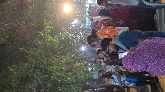 A group of people gathers outdoors at dusk. A few are seated on benches. Some are standing, focused on a digital screen, likely a tablet, and are watching content together. One person wears a purple floral dress. Others are in casual clothes, like jeans. Trees and foliage surround the group, and a streetlamp illuminates the scene. The image appears to be captured from an elevated vantage point.