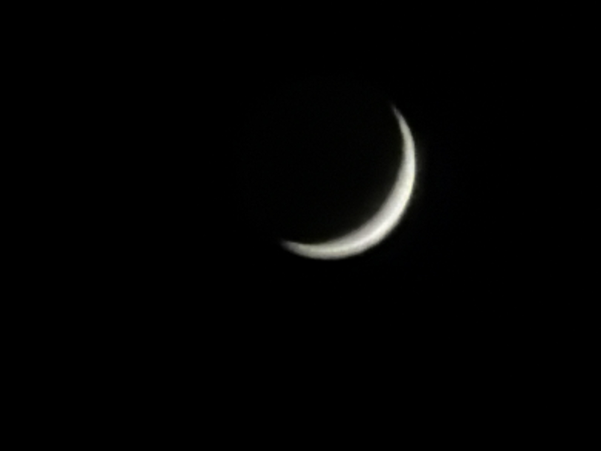 A thin crescent moon is visible in the dark night sky. The moon is illuminated from the right, casting a soft glow. The background is completely black, indicating a clear night with no other visible celestial bodies or light sources. The image appears to be taken from Leudelange, Luxembourg, though no specific landmarks or urban elements are discernible in the frame. The focus is solely on the celestial phenomenon.