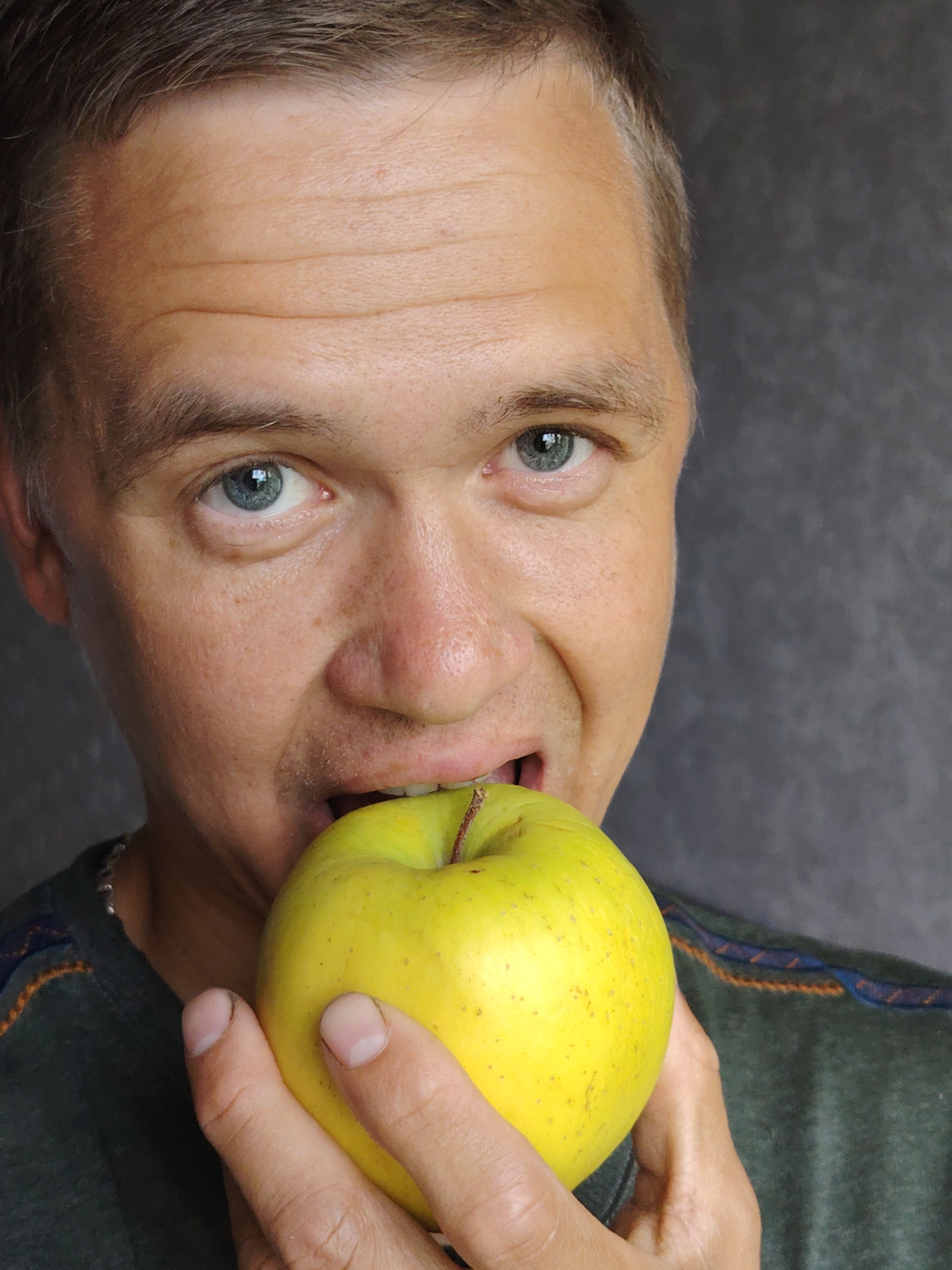 The image is a medium close-up portrait of a light-skinned adult male with short brown hair and blue eyes, who appears to be in his late 30s or early 40s. He is biting into a bright yellow apple. The man is wearing a dark green t-shirt with a thin striped pattern around the neckline. His skin has a few freckles and some visible pores. The background is a plain, dark gray textured surface. The lighting suggests an indoor setting, possibly with artificial light. There are no specific identifiable landmarks or context to place this image within the city of Chaikovsky, Russia.