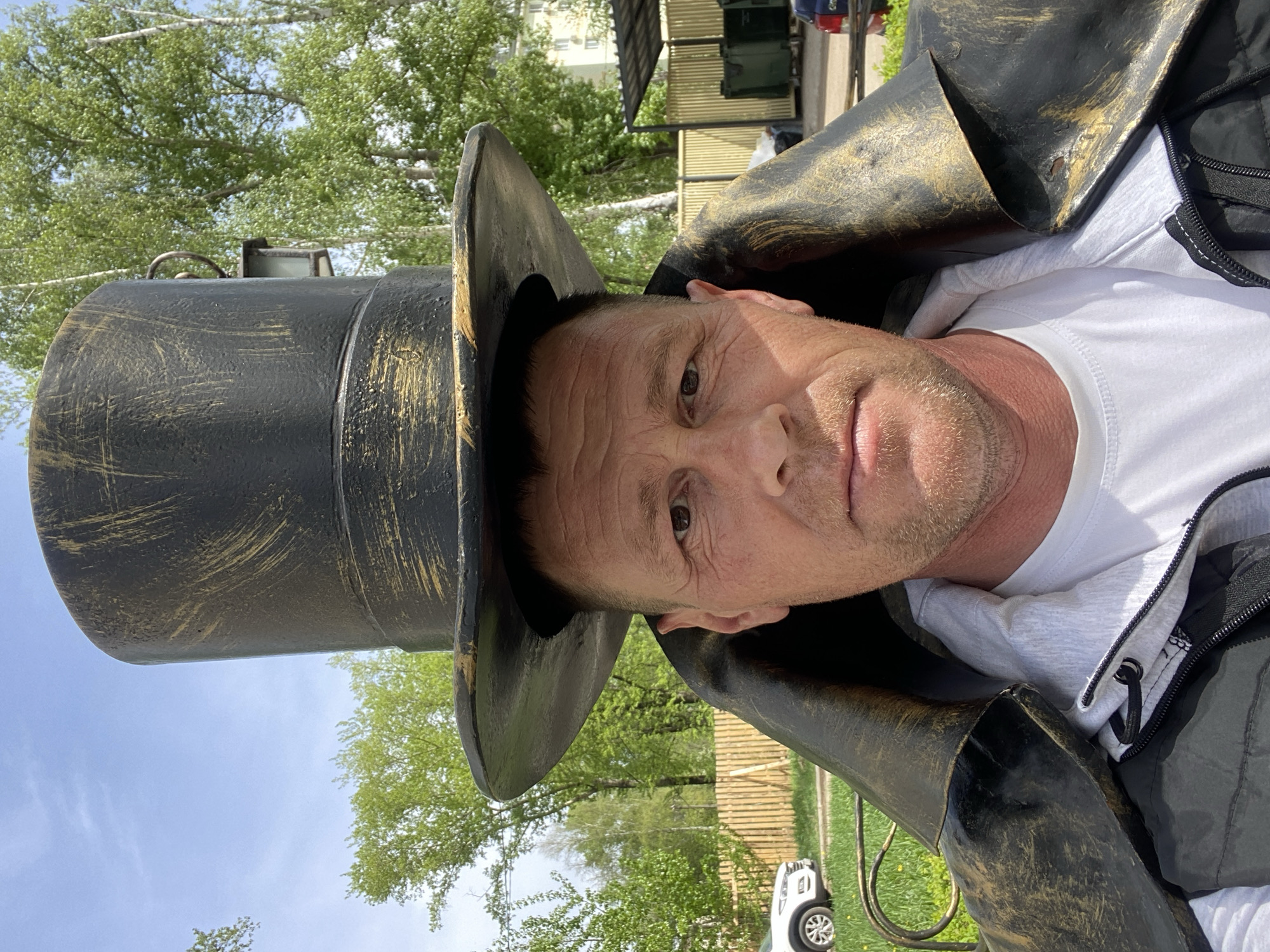 A man wearing a black and gold top hat is posing for a selfie outdoors. He is also wearing a white t-shirt and a dark jacket. The location is likely Chaikovsky, Russia, given the context. In the background, there are green trees, a wooden fence, a white car, and a building with green siding. The sky is partly cloudy. The image is rotated counter-clockwise.