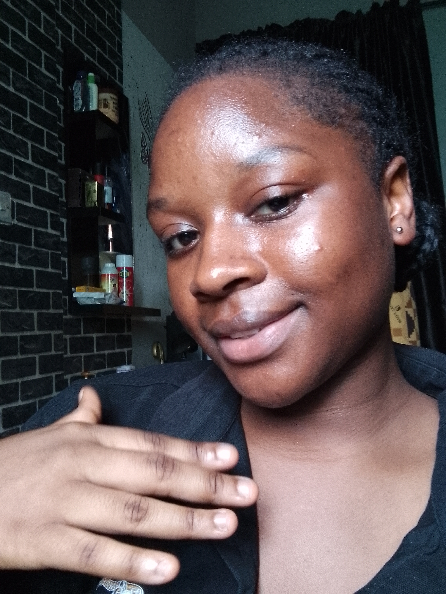 A close-up, eye-level indoor shot features a dark-skinned individual, likely female, with short, dark hair, looking towards the camera with a subtle smile. Her skin appears moisturized or slightly dewy. She wears a dark, possibly black, collared shirt. Her left hand is raised, partially covering her lower face and chest, revealing a small decorative item or ring on her pinky finger. A small silver stud earring is visible in her left earlobe.
The background consists of a dark, brick-patterned wall on the left. Mounted on this wall are dark, multi-tiered shelves. The top shelf holds various bottles and containers, including one that is light green/white and another brown container. The middle shelf displays more items, such as a white/red spray can, a yellow/white bottle, and small boxes. A faint light source or small flame is visible on this shelf. To the right, a dark curtain hangs, and a lighter-colored wall with faint abstract markings is visible. The lighting is dim, suggesting an indoor setting. No discernible text is clearly readable on any of the products.