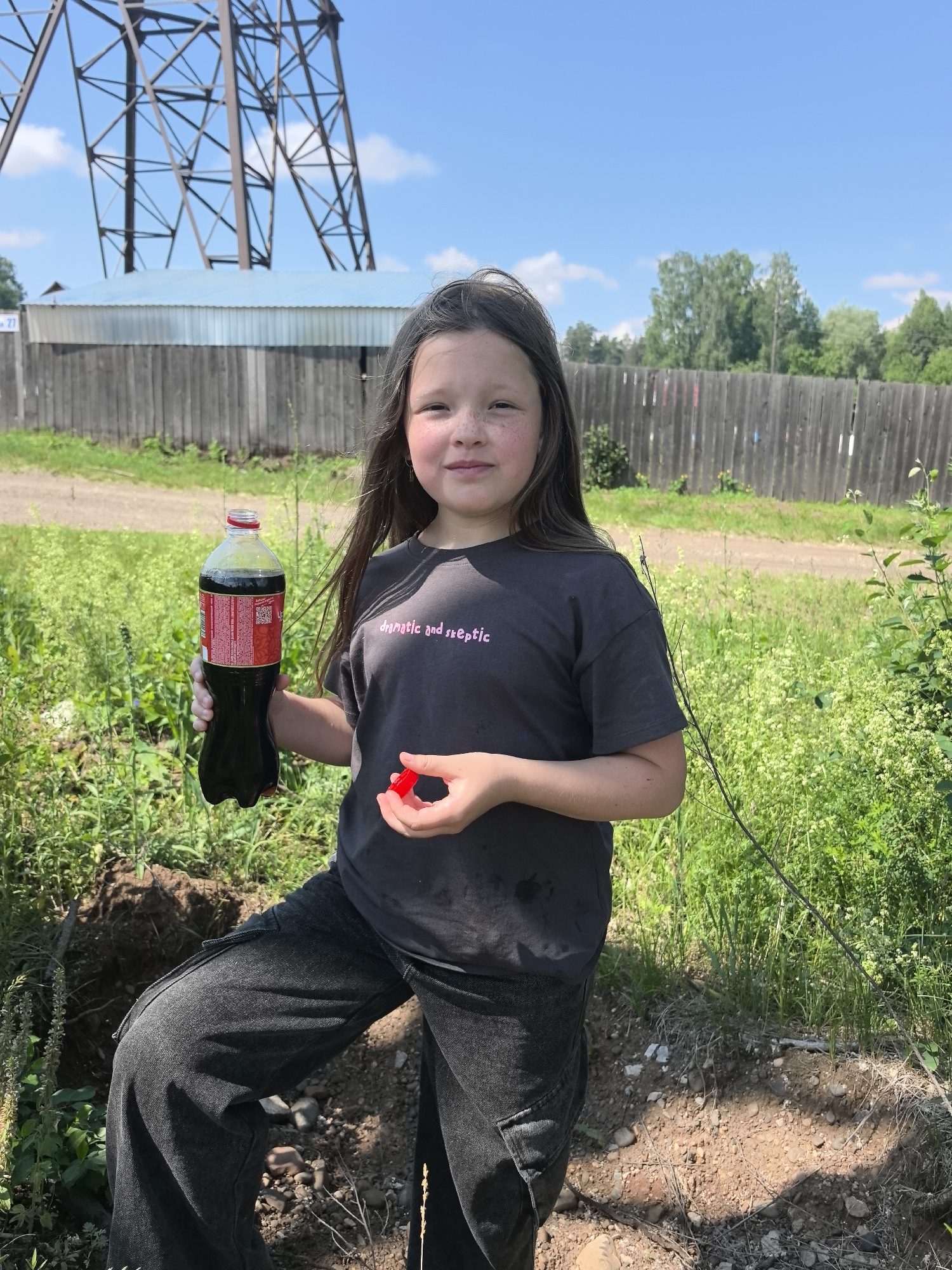 A young girl with long, dark hair and freckles is standing outdoors, holding a bottle of Coca-Cola in her left hand and a red cap in her right. She is wearing a dark gray t-shirt with pink text and dark gray baggy pants. The t-shirt has the text "dogmatic and skeptic" printed on it. The girl is smiling at the camera.
She is standing in a grassy area with a dirt path behind her. In the background, there is a wooden fence and a large metal structure, possibly a transmission tower. There are trees and greenery visible, and the sky is blue with some scattered clouds. The lighting suggests it is daytime, likely late morning or early afternoon. The overall scene appears to be rural, consistent with the provided location context of Noviy, Russia. The activity seems casual, with the girl posing for a picture while holding the drink.