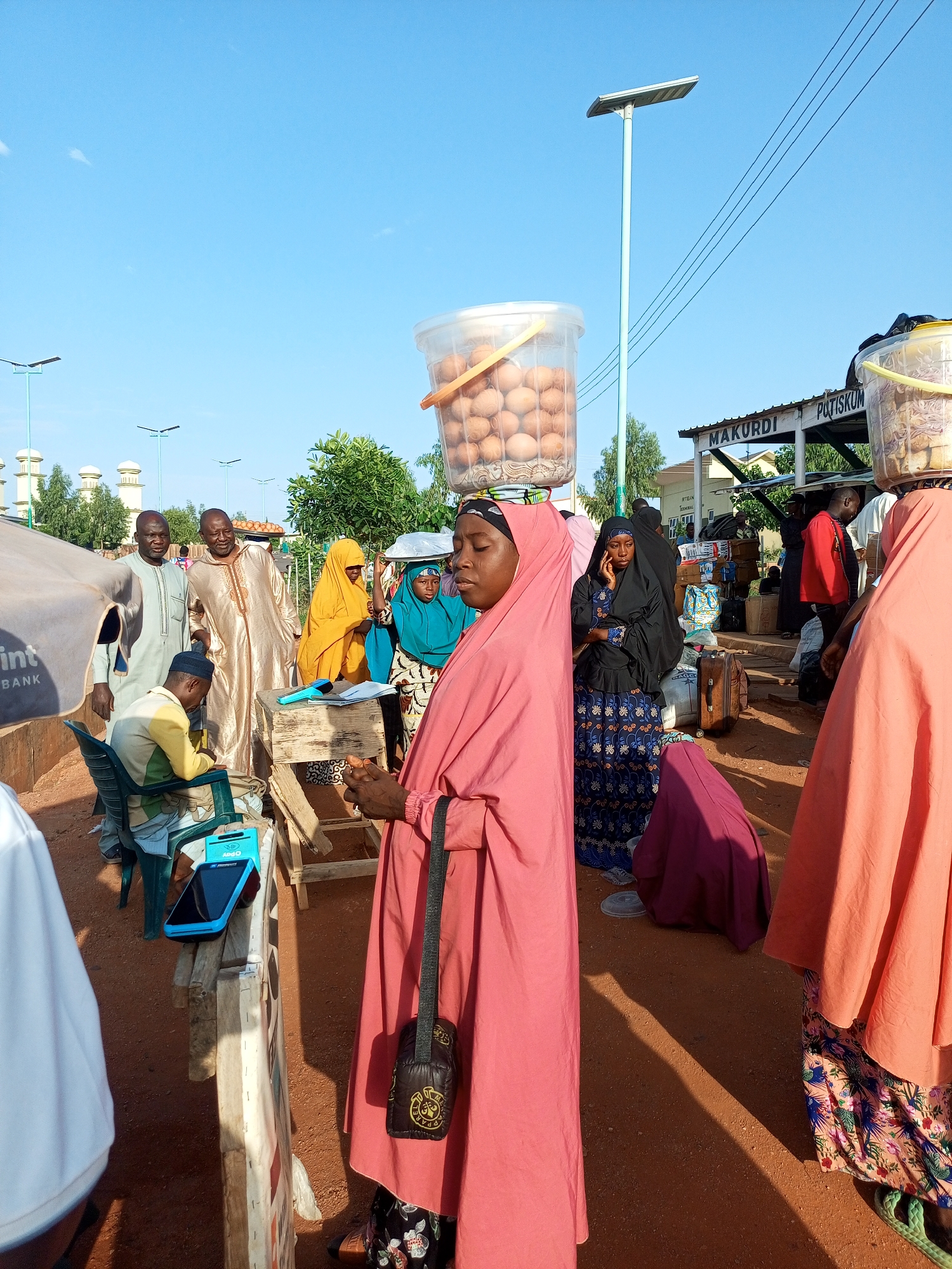In the city of Gombe, Nigeria, this outdoor marketplace scene captures a bustling atmosphere with several individuals engaged in commerce. A woman in a vibrant pink hijab and dress stands in the foreground, her gaze directed downwards. Behind her, another woman in a black hijab and patterned blue dress is on her phone.
The marketplace is characterized by dirt ground and several stalls or stands. One stand features a wooden structure with a transaction device, possibly a point-of-sale terminal, and a stack of papers. To the left of this stand, a young man in a yellow and white checkered shirt and cap is seated, holding a blue electronic device.
Several people are seen in traditional attire, including men in flowing robes and women in hijabs of various colors, such as yellow and green. Some individuals are carrying containers of goods on their heads; one woman has a large clear plastic bucket filled with eggs. Another person is seen carrying what appears to be a tray on their head.
In the background, a structure with "MAKURDI POTISKUM" is visible, likely indicating a transport hub or a specific area within the market. Multiple lampposts with solar panels are present, suggesting an effort towards modernization in the public lighting infrastructure. The sky is a clear blue, indicating a sunny day. Various bags and luggage are scattered around, suggesting travel or goods being transported. The overall impression is one of an active and vibrant local marketplace.