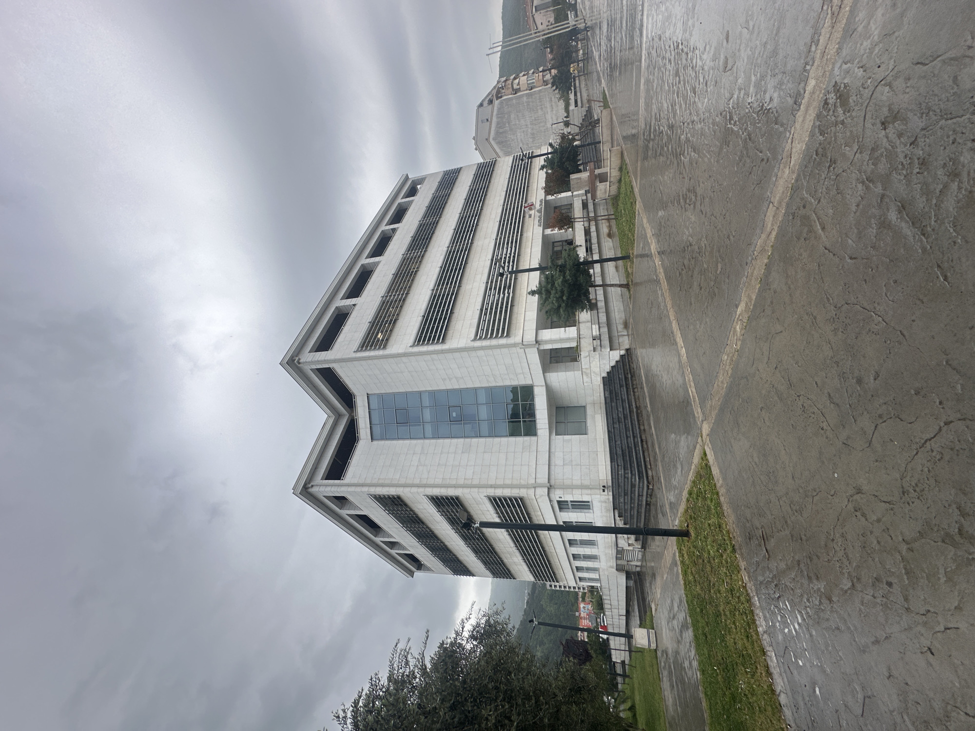 This media file captures a modern building in Budva, Montenegro, under an overcast sky. The image is taken from a slightly low angle, looking towards the building, which dominates the left side of the frame.
The main subject is a multi-story, light-colored (likely white or grey stone/concrete) building with contemporary architecture. It features a distinctive V-shaped cut-out at its top center. Large rectangular windows, some tinted and reflecting the sky, are visible, along with horizontal louvers or shading elements on several facades. Entrances to the building are visible on the lower levels, flanked by streetlights.
The ground in the foreground and leading up to the building is a wet, dark grey paved area, possibly stamped concrete, with lighter grey lines creating a grid pattern. The wet surface suggests recent rain. Patches of green grass are present between the paved areas and around trees. Scattered dark green trees and bushes are seen in the foreground and near the building.
In the mid-ground and background, other buildings can be discerned, with one appearing unfinished or under construction. Distant hills or mountains, hazy and partially obscured by clouds, form the backdrop.
The sky is uniformly overcast and grey, indicating a cloudy or rainy day. The overall lighting is diffuse, without direct sunlight. There are no people visible in the image.
On the main building, near one of the entrances, a sign is visible with a red circular logo. Below the logo, text in Cyrillic script can be partially made out, including "Агенција" (Agency) and "ЦЕНТАР" (Center). Another red banner with white text is visible on a lamppost further down the street, but its text is illegible due to distance.